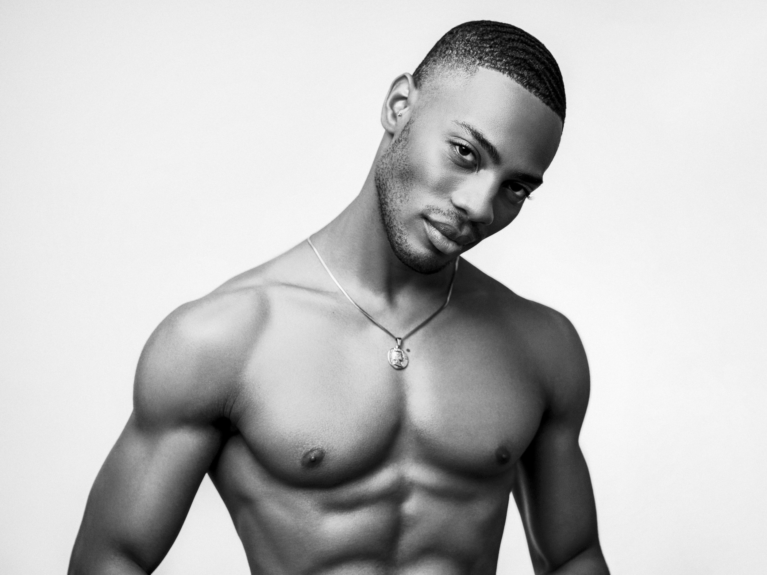 Black and white photo of a shirtless young man with styled hair, a necklace, and a confident expression, posing against a plain background.