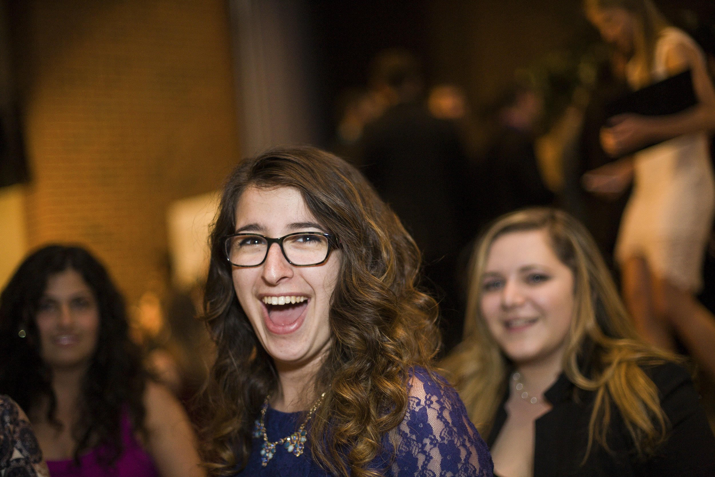 Young woman with glasses and curly hair smiling at a social event, surrounded by other women in an indoor setting.