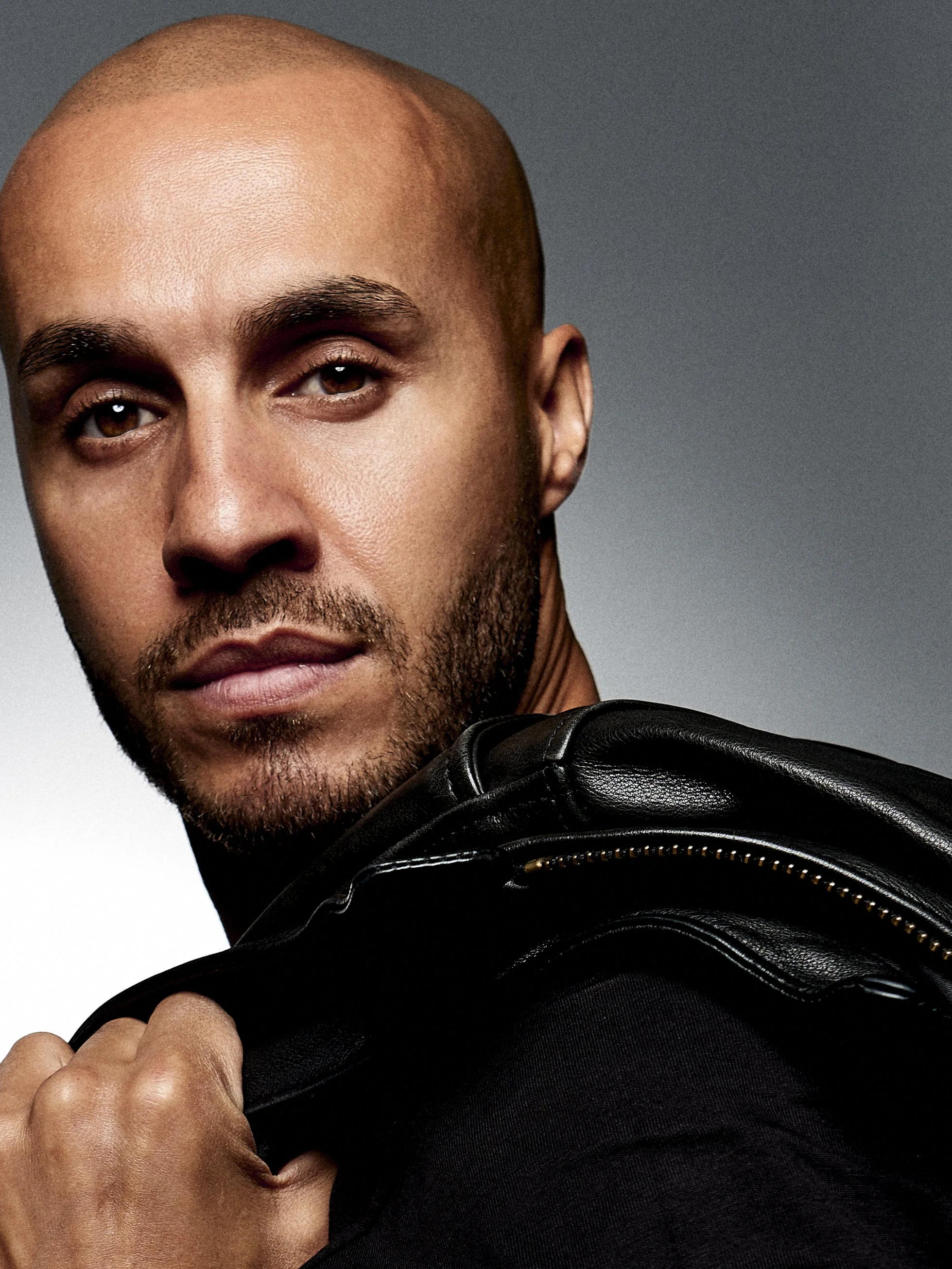 Close-up of a man with a bald head, beard, and mustache, wearing a black leather jacket, against a gray background.