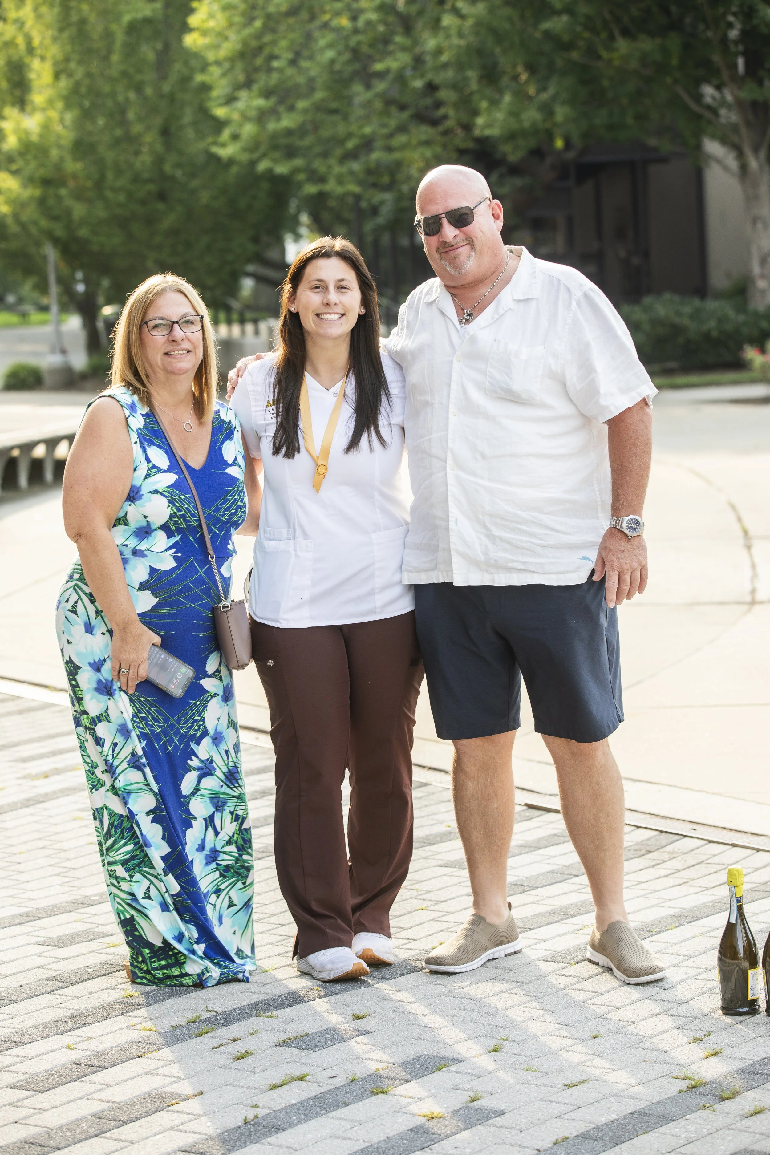 A group of three people standing outdoors on a sunny day, smiling, with trees and a walkway in the background. The woman on the left is wearing a colorful floral dress and glasses, the woman in the middle is wearing medical scrubs and a medal around 