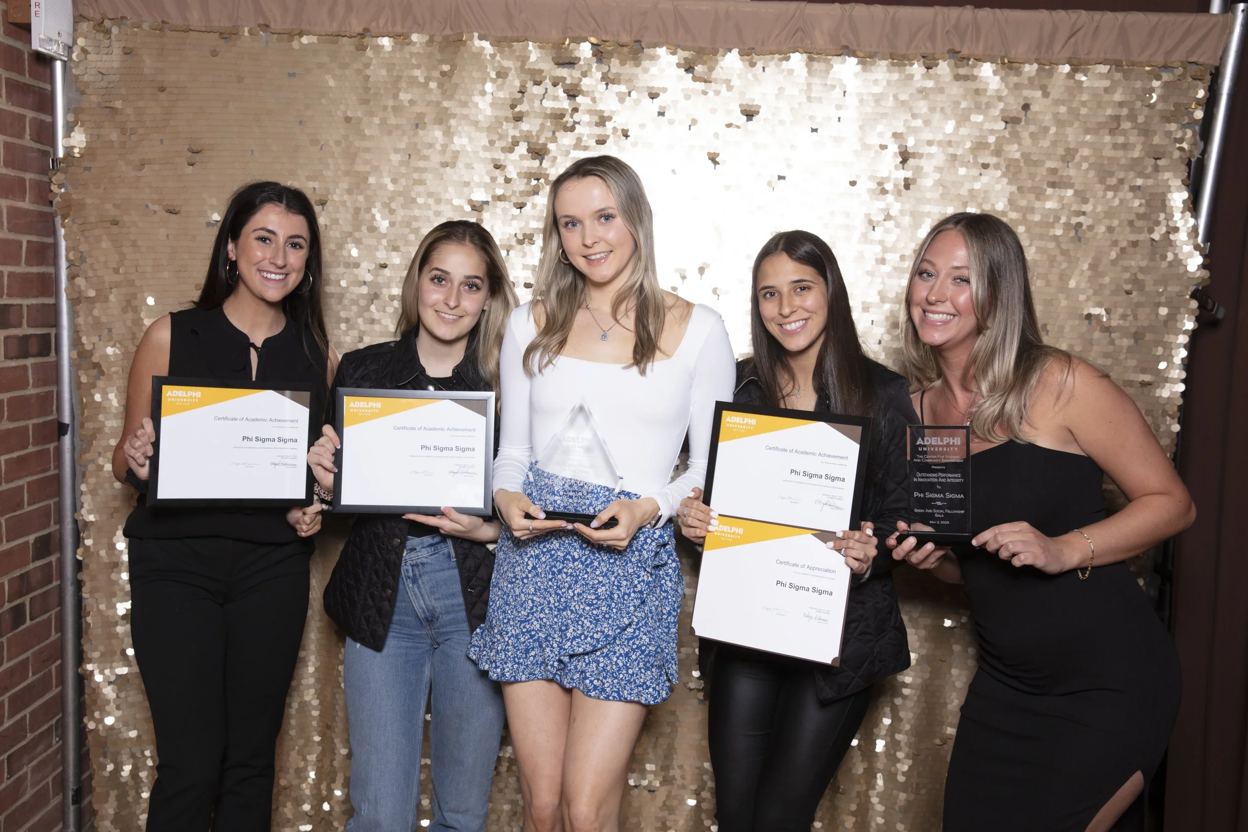 Five young women standing together at an awards ceremony, holding certificates and a glass plaque, in front of a gold sequin backdrop.