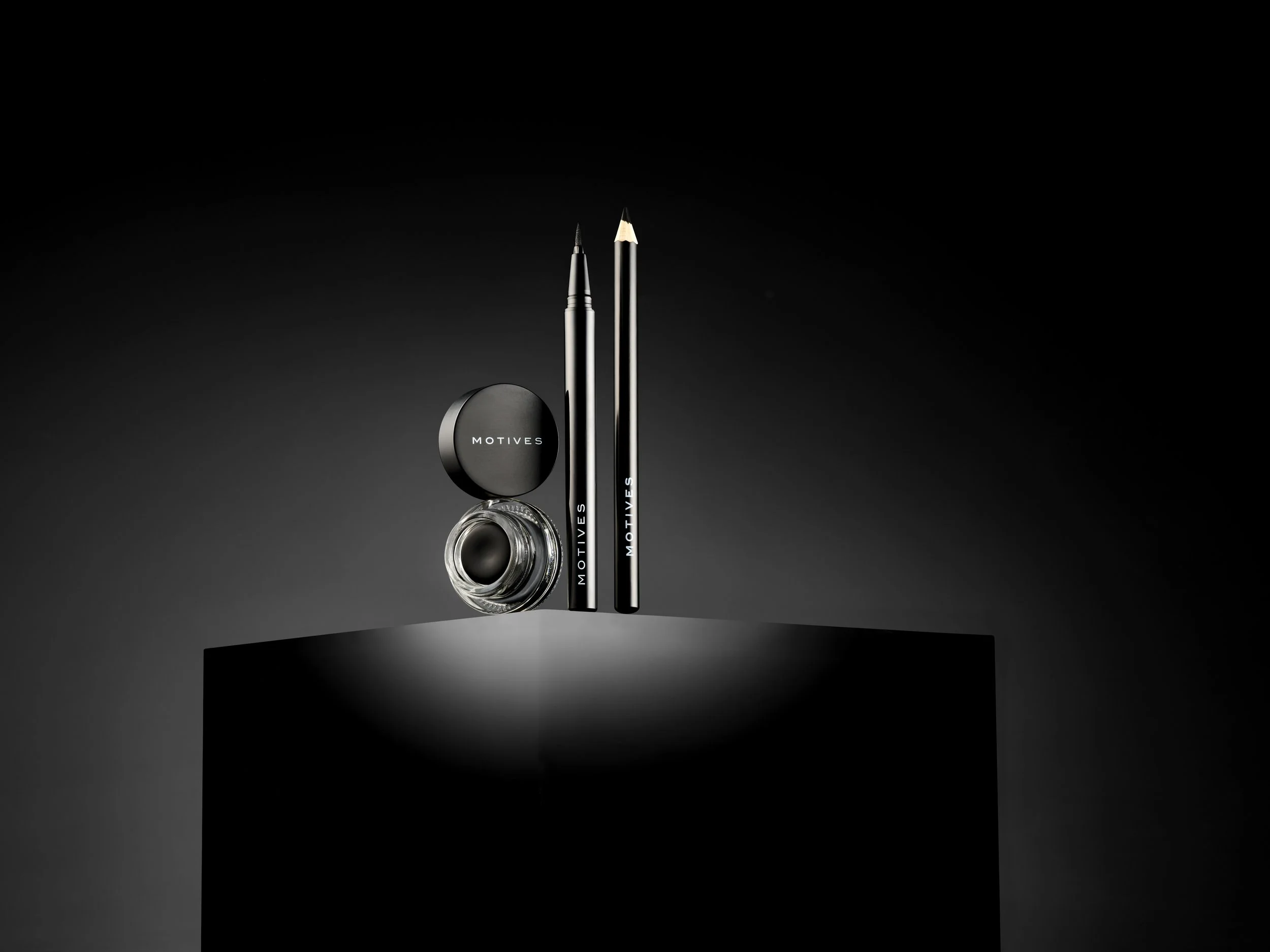 Black and silver eyeliner, brush, and lid container with 'MOTIVES' branding on a black pedestal in a dark background.