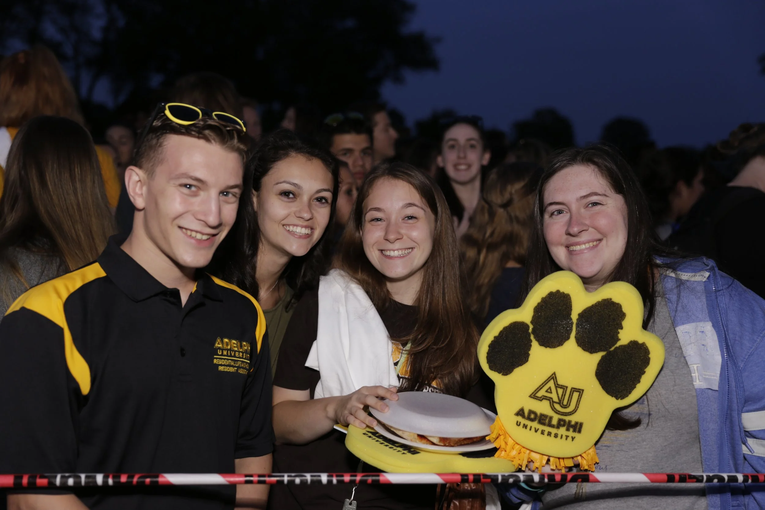 Group of four young people smiling at night, one holding a foam paw and a paper plate with food, wearing clothing and accessories with Auburn University and Adelphi University logos, in a crowd.