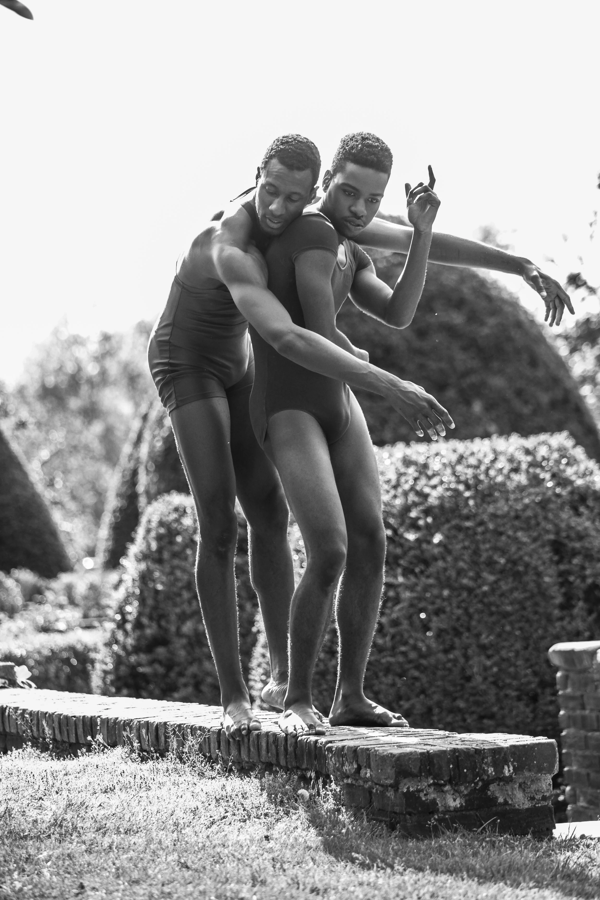 Two male athletes balancing on a narrow brick ledge outdoors, one assisting the other in a coordinated pose in black and white.
