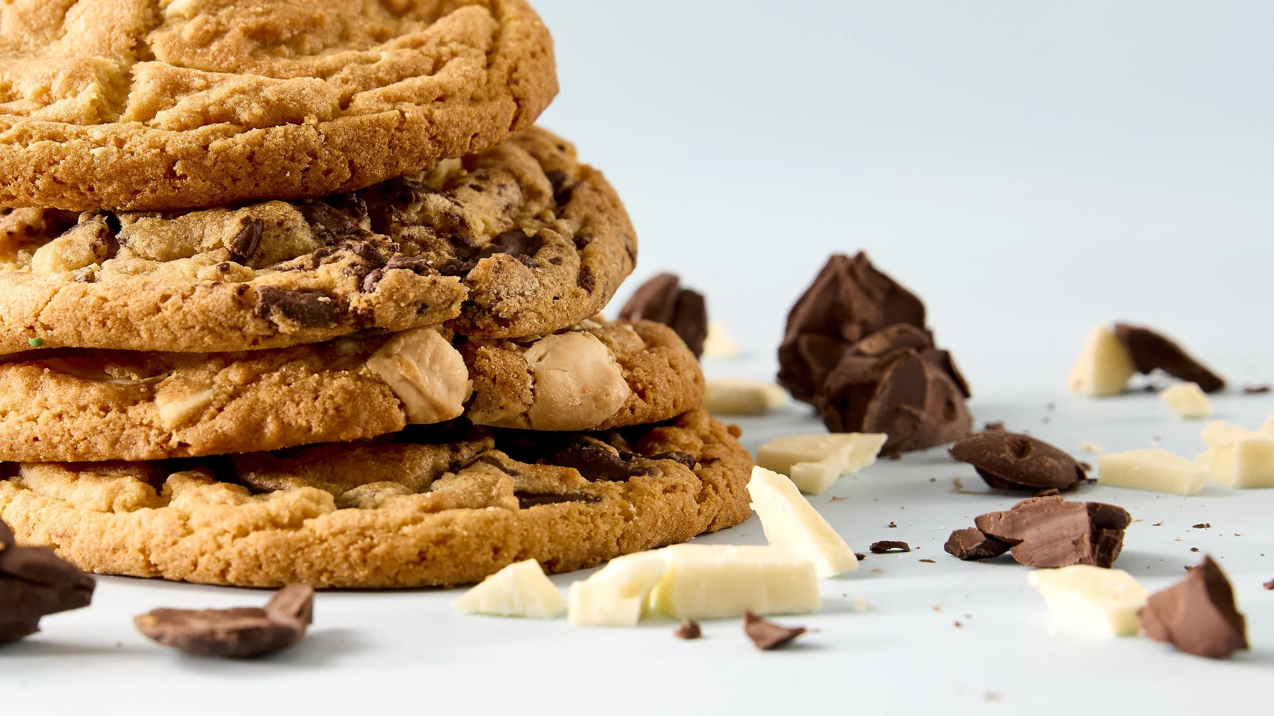 Close-up of three large cookies stacked on top of each other, with chunks of chocolate and white chocolate chips visible. Scattered pieces of chopped white and dark chocolate surround the cookies on a light-colored surface.