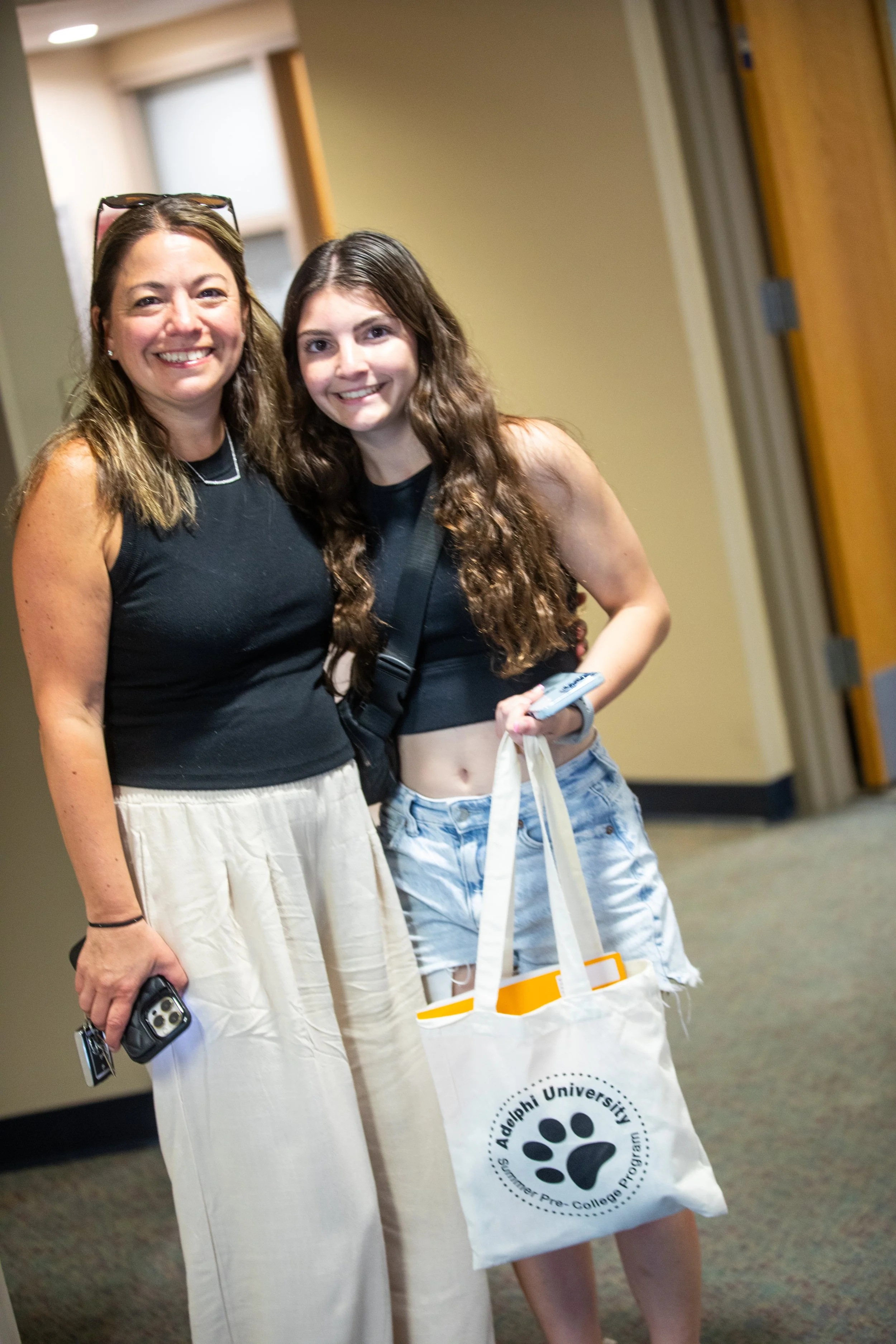 Two smiling women standing indoors, one holding a smartphone and a tote bag with the Adélphi University logo, in a hallway with beige walls and a wooden door.