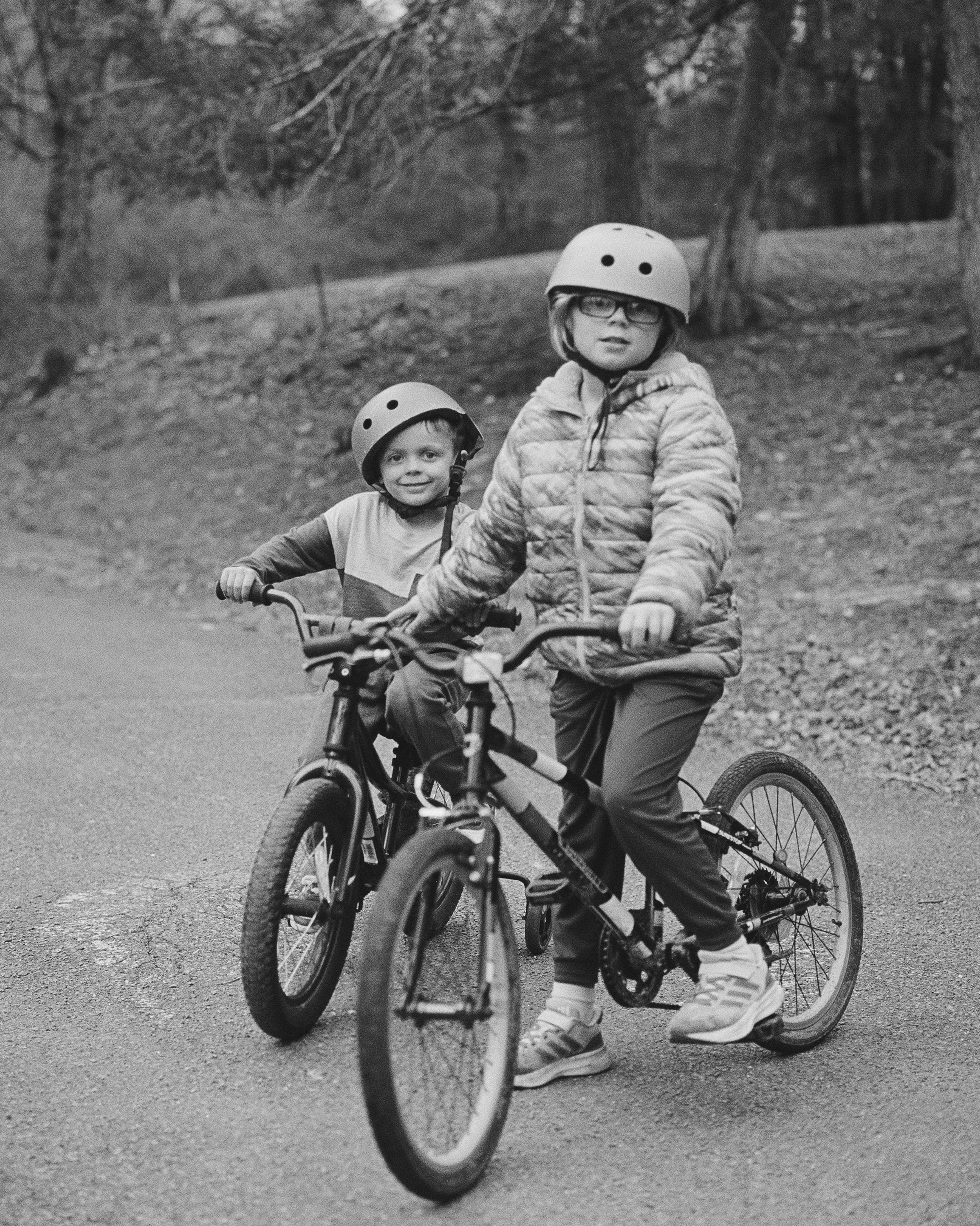 Two children riding bicycles outdoors, wearing helmets and jackets, in a park or wooded area.