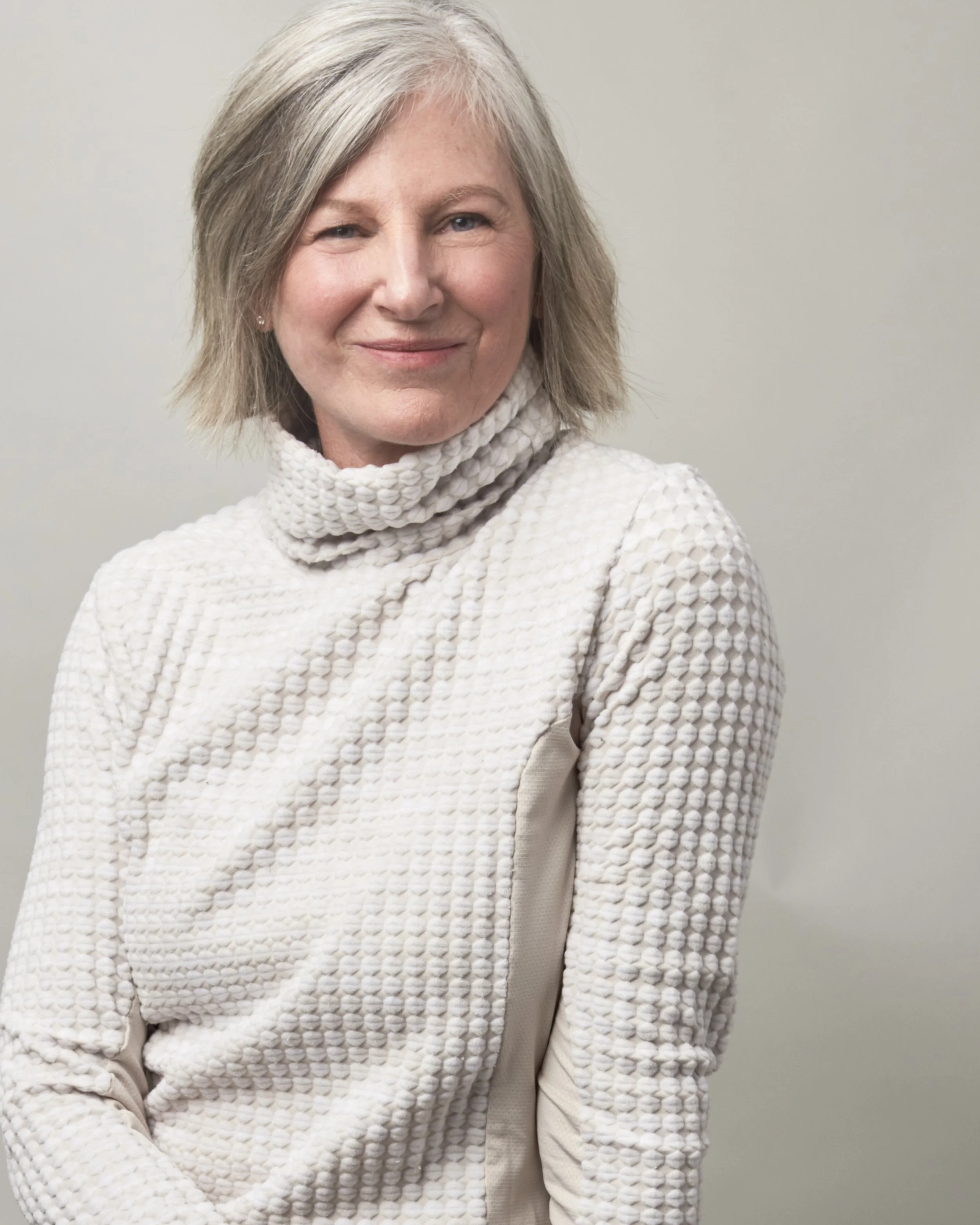 A woman with shoulder-length gray hair smiling, standing against a plain white background, wearing a textured cream-colored turtleneck sweater.