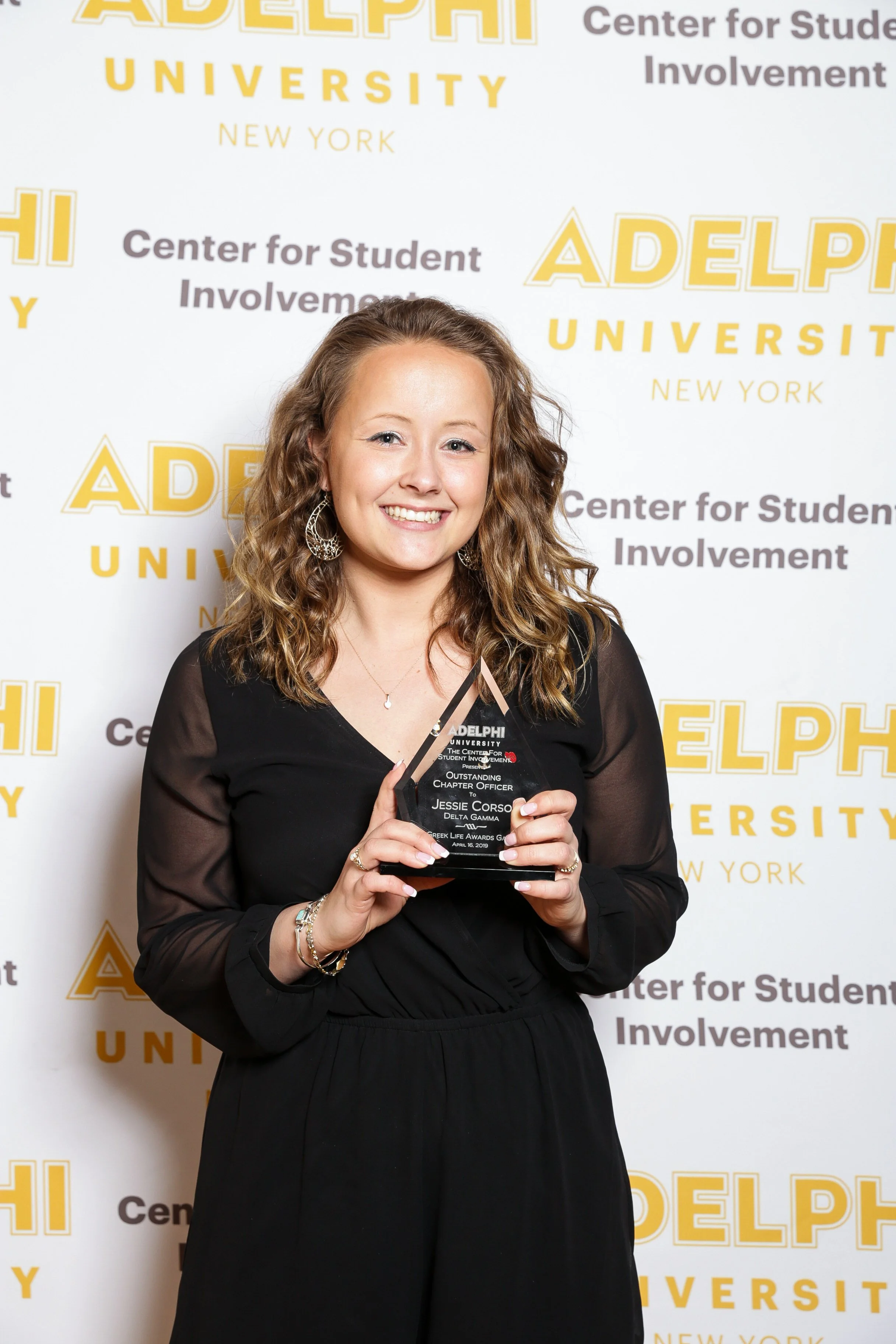 A young woman with curly brown hair smiling while holding a glass award in front of a backdrop with Adelphi University and Center for Student Involvement logos.