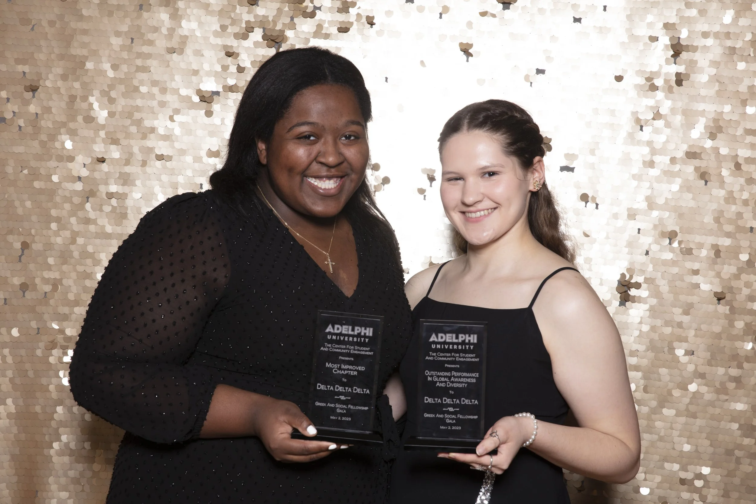 Two women smiling and holding awards at a formal event with a gold sequin backdrop. The woman on the left is wearing a black dress with sheer sleeves and a cross necklace. The woman on the right is wearing a black spaghetti strap dress with pearl ear