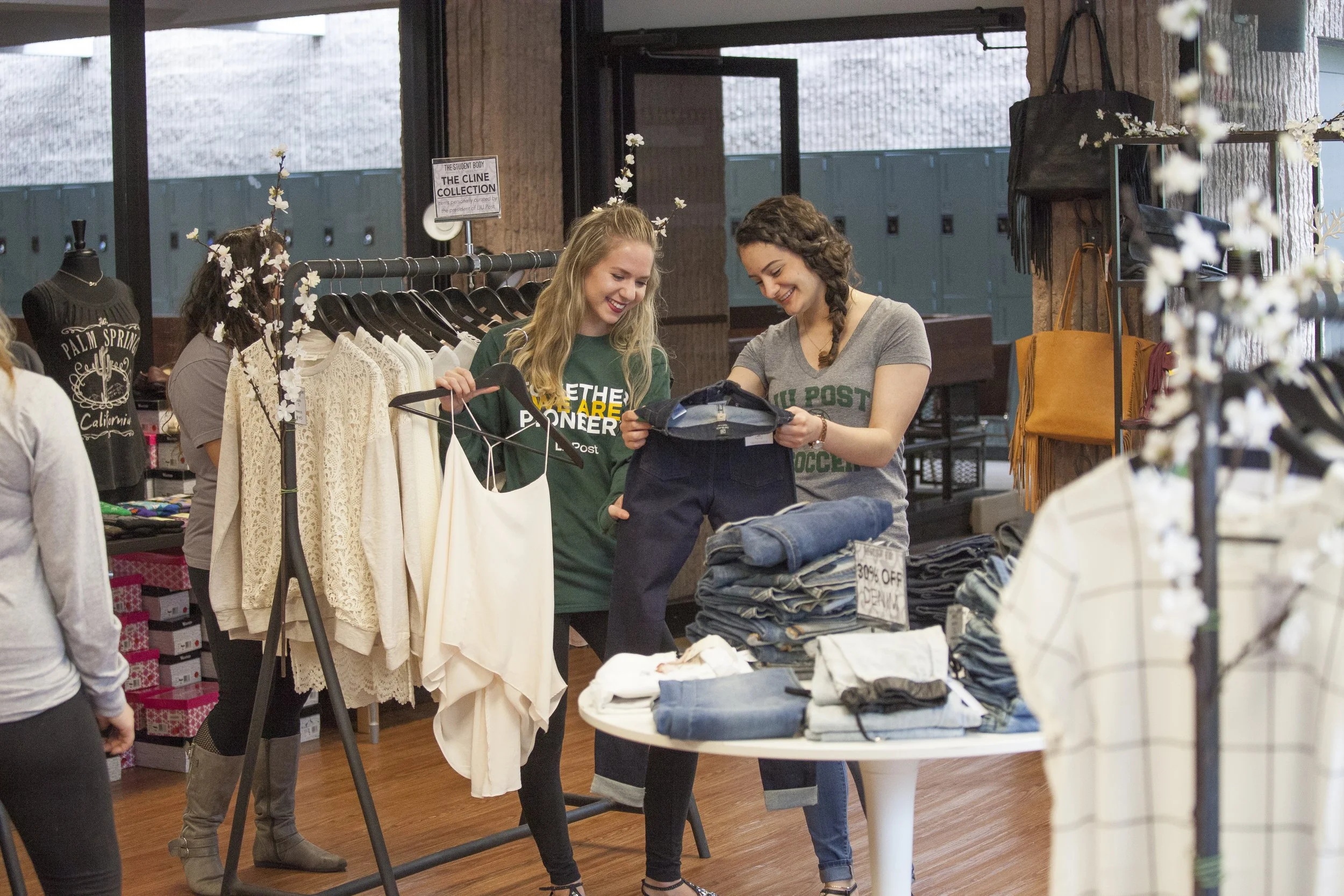 Two young women shopping for clothes inside a retail store, smiling and holding clothing items.