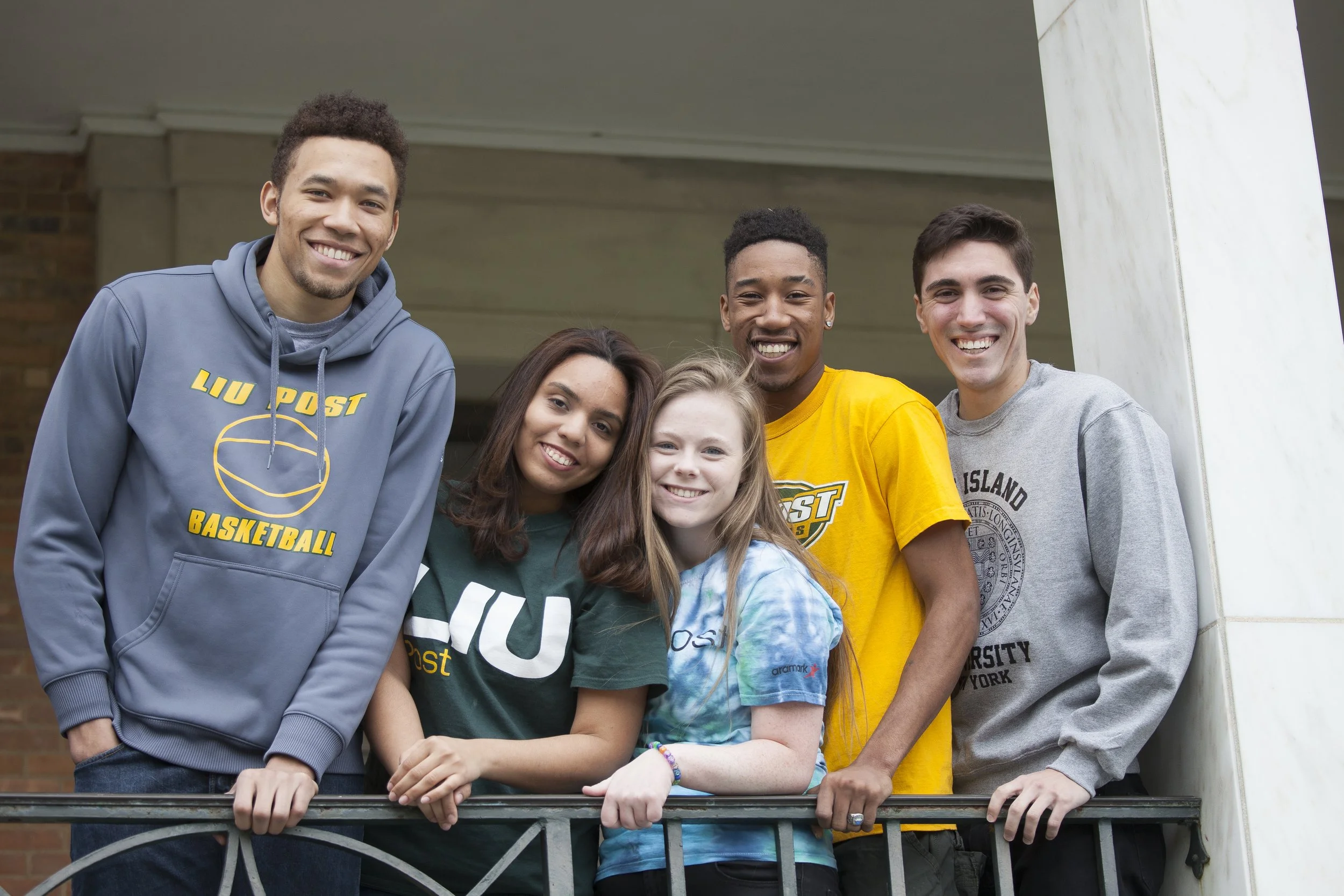 Group of six diverse young people smiling on a balcony with brick and marble in the background.