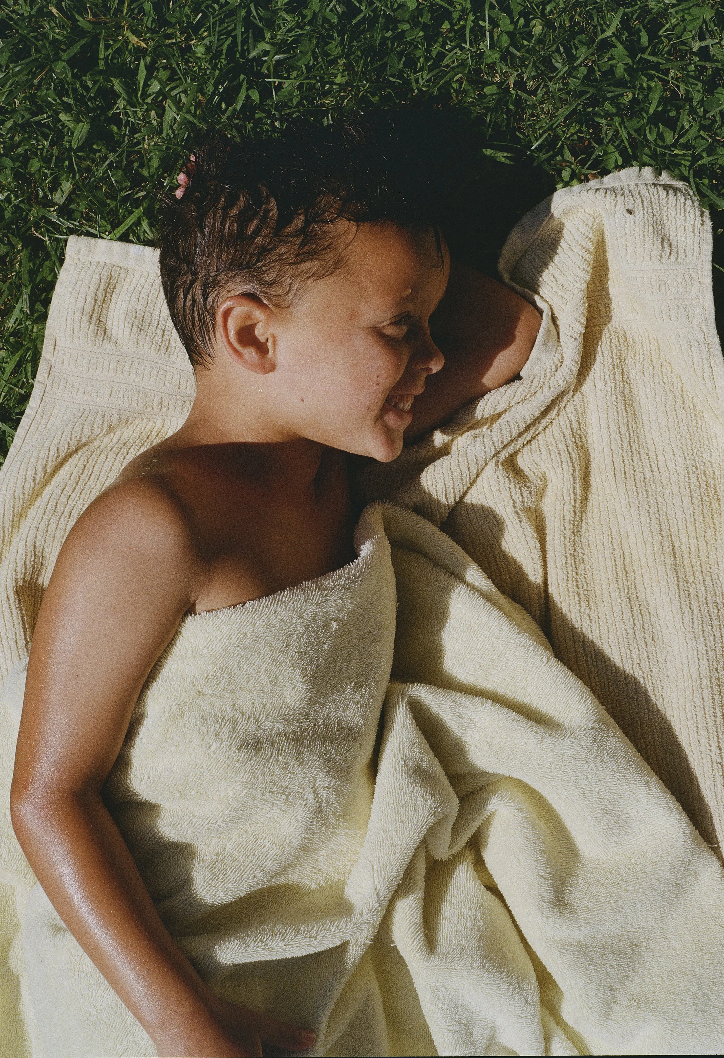 A person with wet hair smiling and lying on a towel in grass.