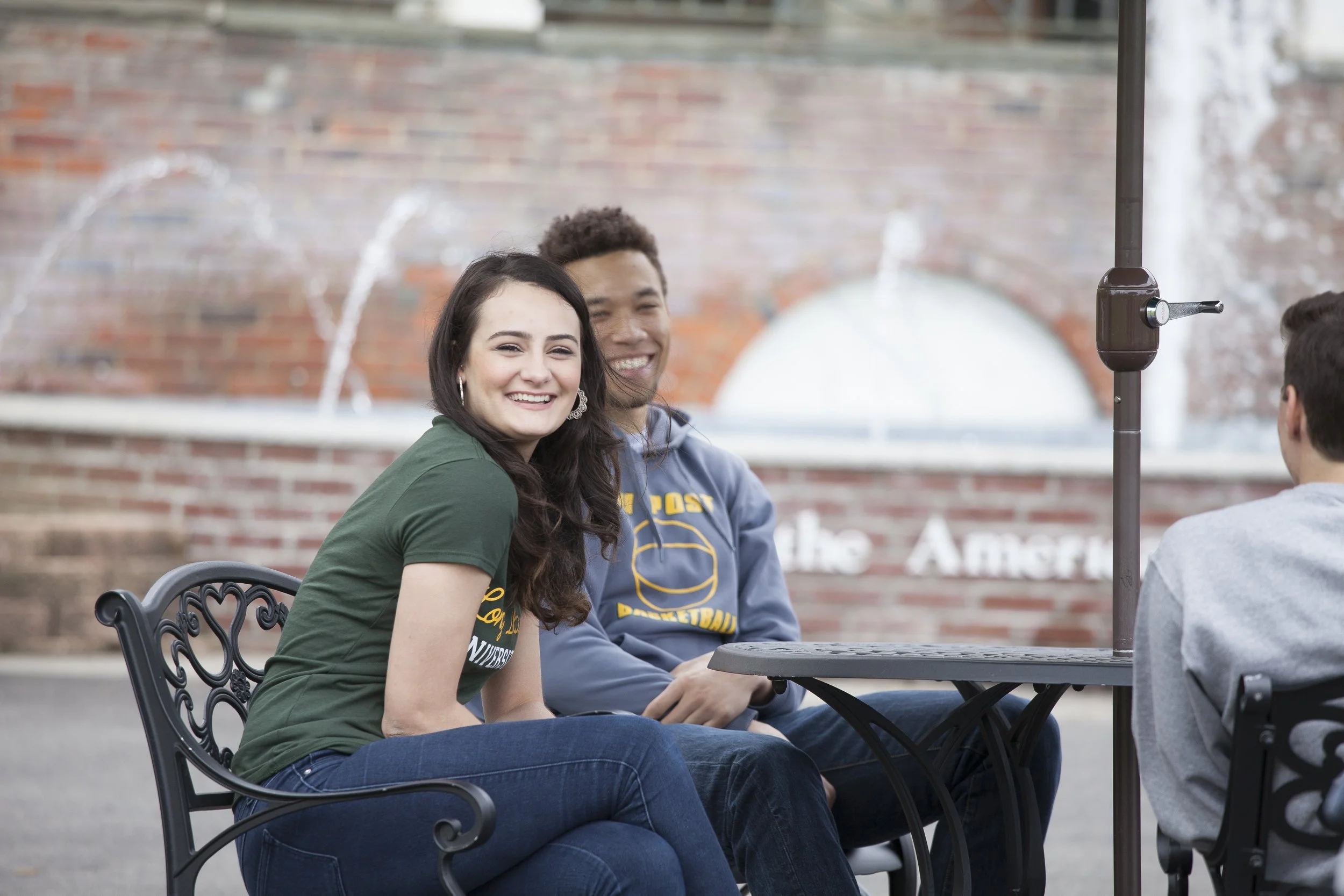A group of young people sitting at a table outdoors, smiling and enjoying each other's company, with a brick wall and fountains in the background.