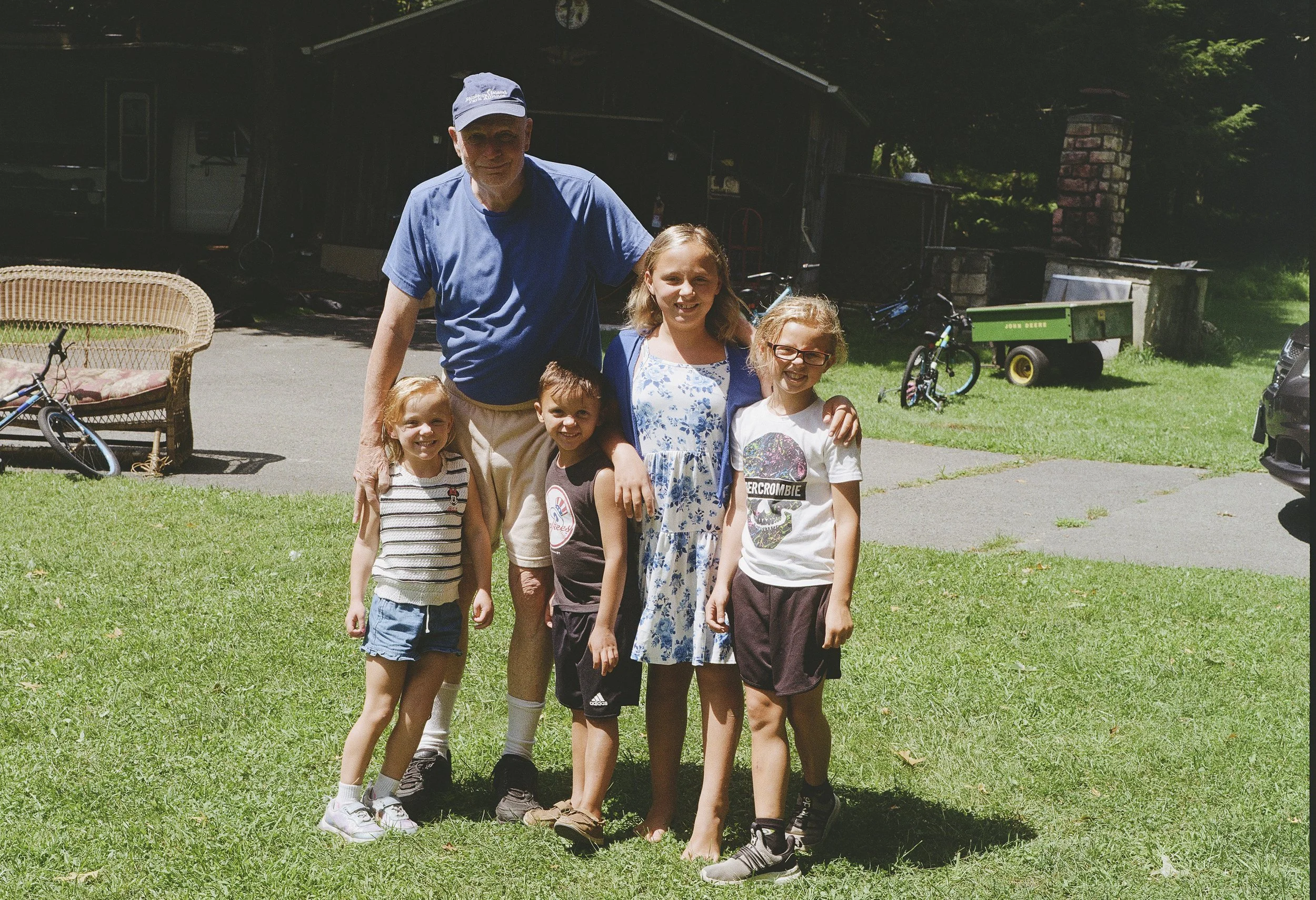 A group of six children and one adult posing outside on a grassy area. The adult is an older man wearing a blue t-shirt and a baseball cap. The children are smiling and standing close together around him, dressed in casual summer clothes. There are b