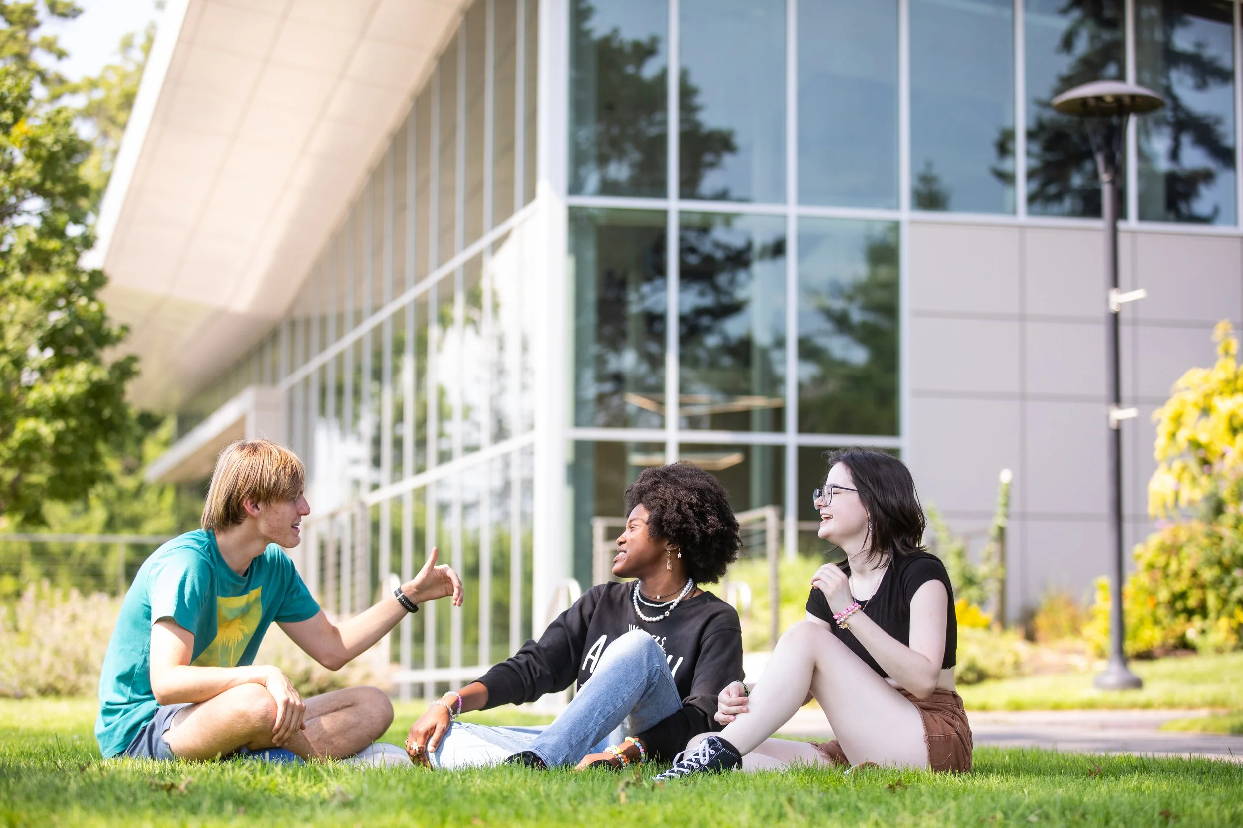 Three young people sitting on grass outdoors talking, with a modern building with large glass windows in the background