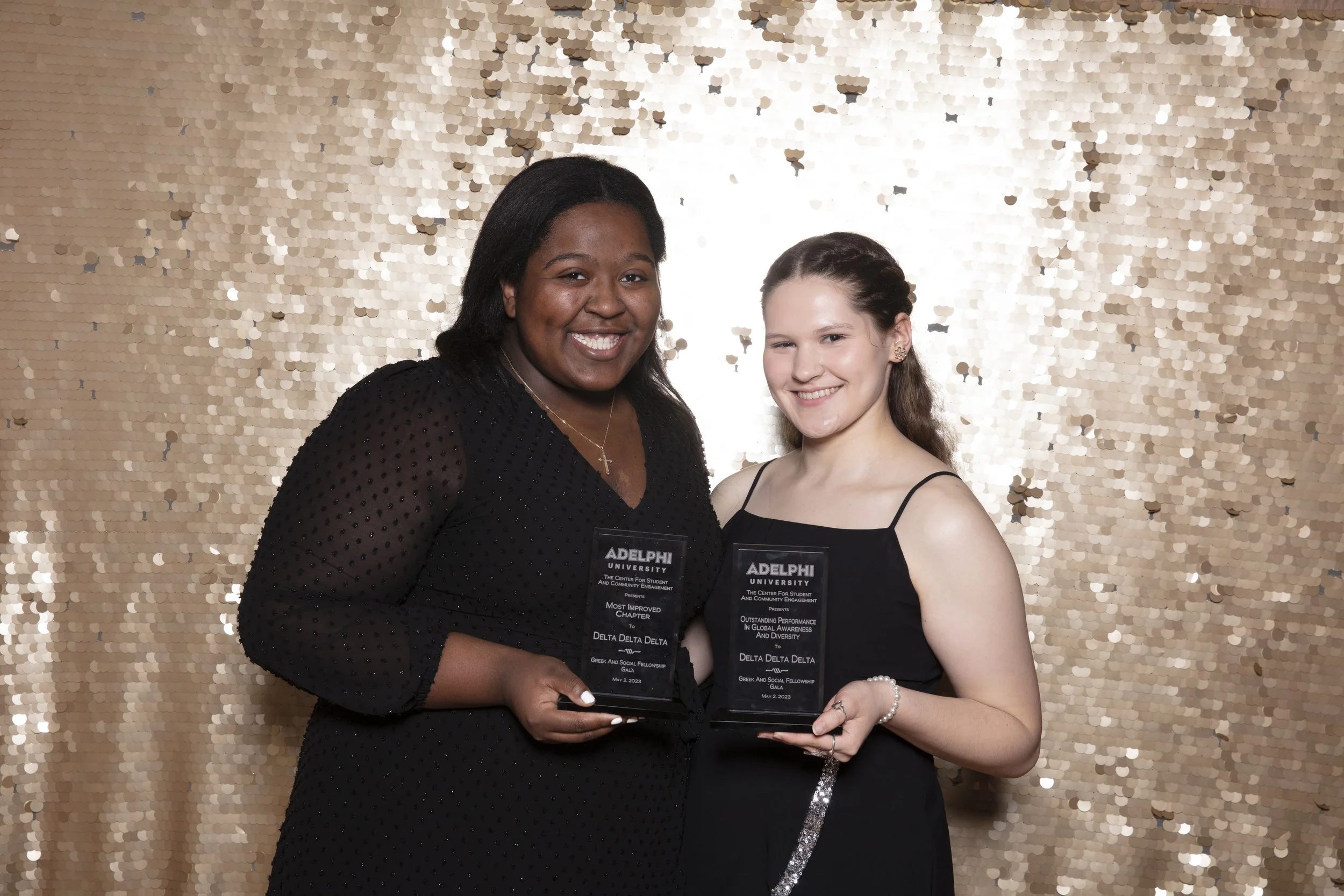 Two women in black dresses holding awards and smiling at a photo event with a gold sequin backdrop.