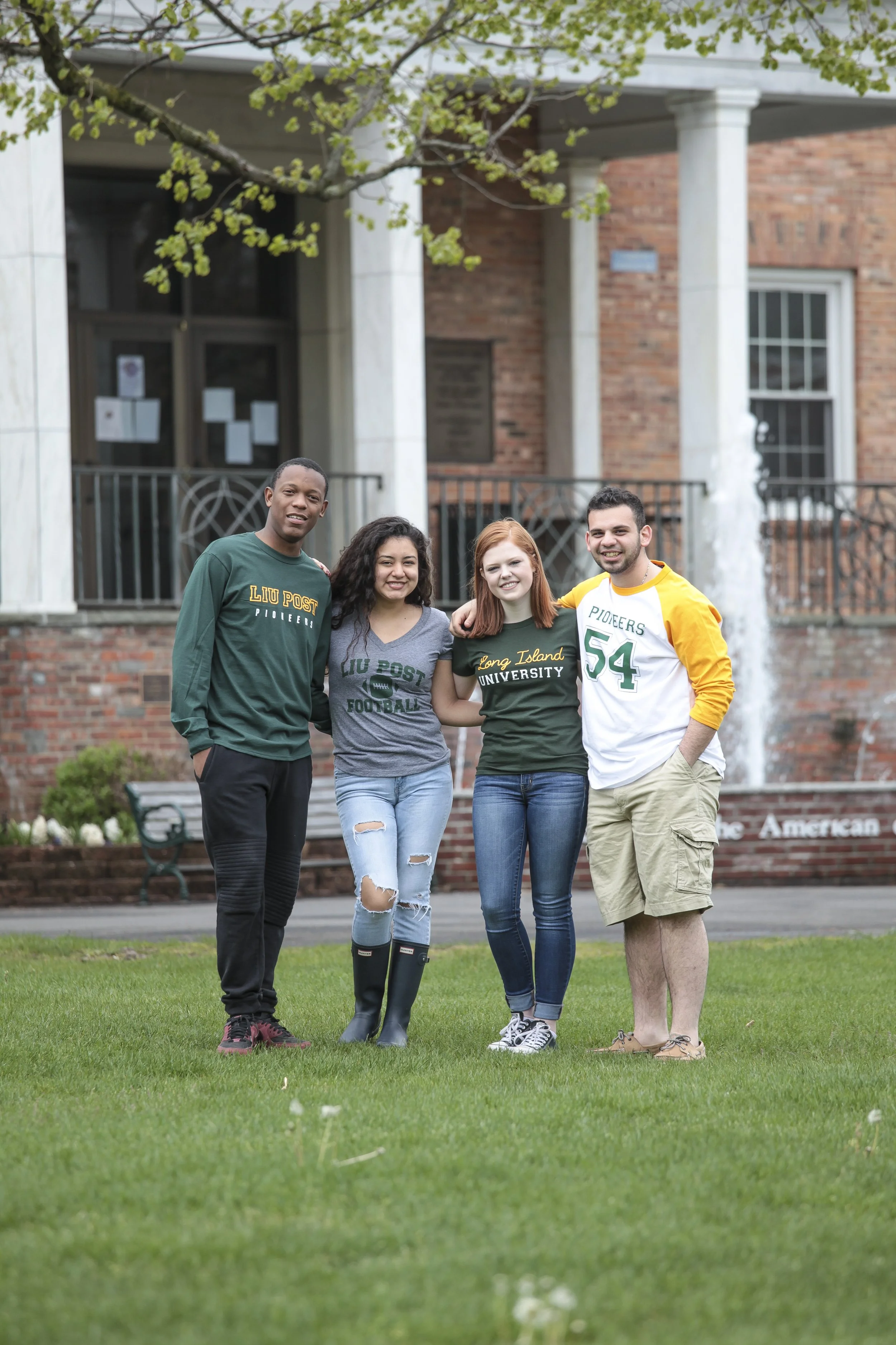 Four young adults standing together outside in a grassy area, smiling, with a brick building and trees in the background.
