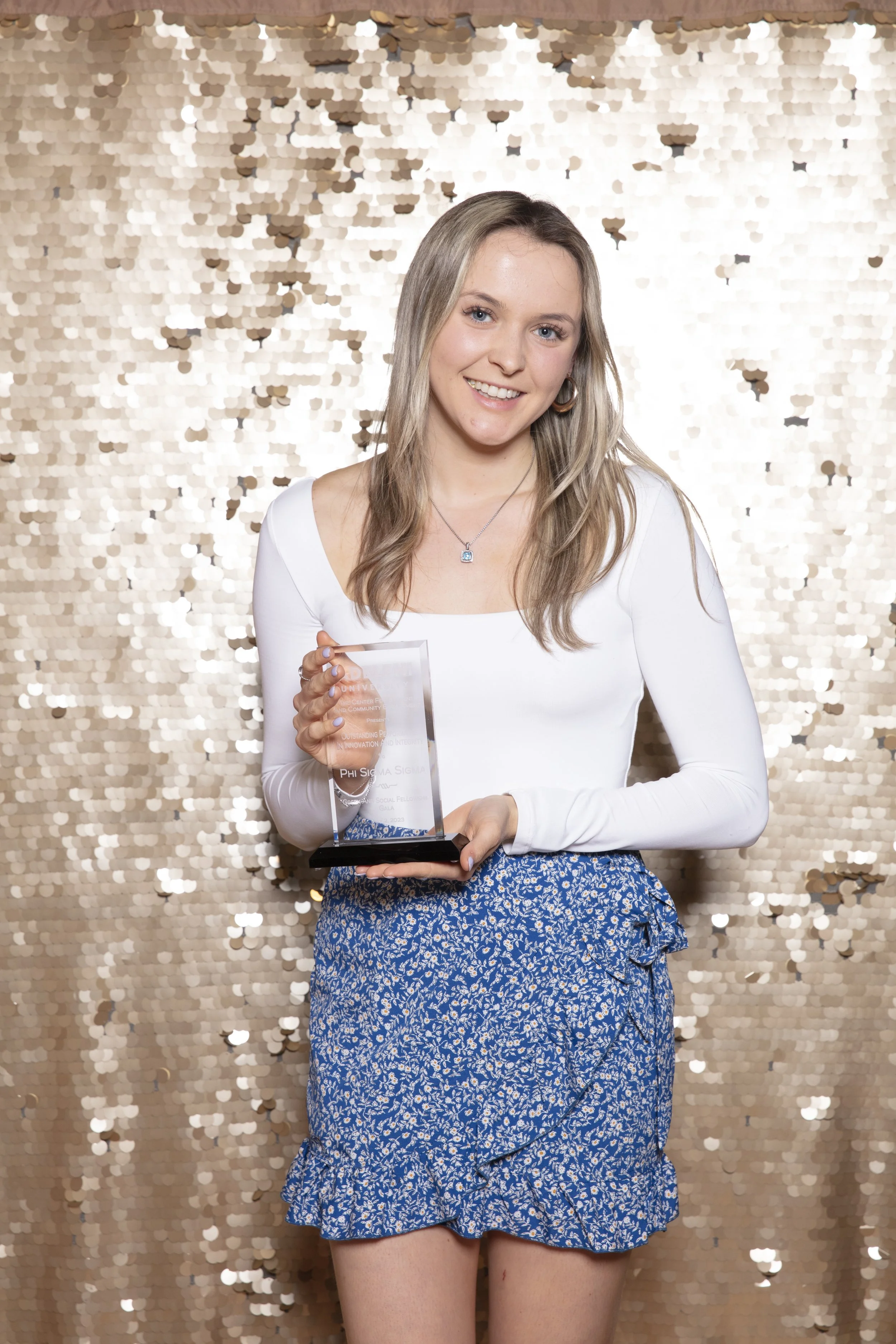 A young woman with long blonde hair wearing a white long sleeve top and blue patterned shorts posing with a clear award against a gold sequin backdrop.
