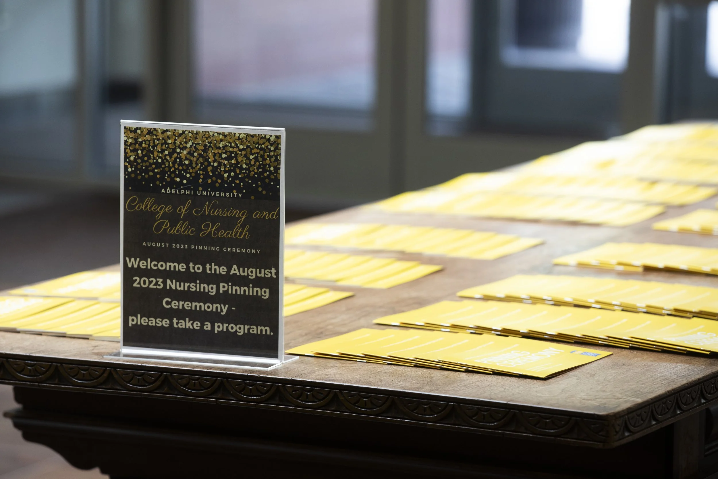 An informational sign on a wooden table at an August 2023 nursing pinning ceremony at Adelphi University, with multiple yellow program booklets arranged on the table in the background.