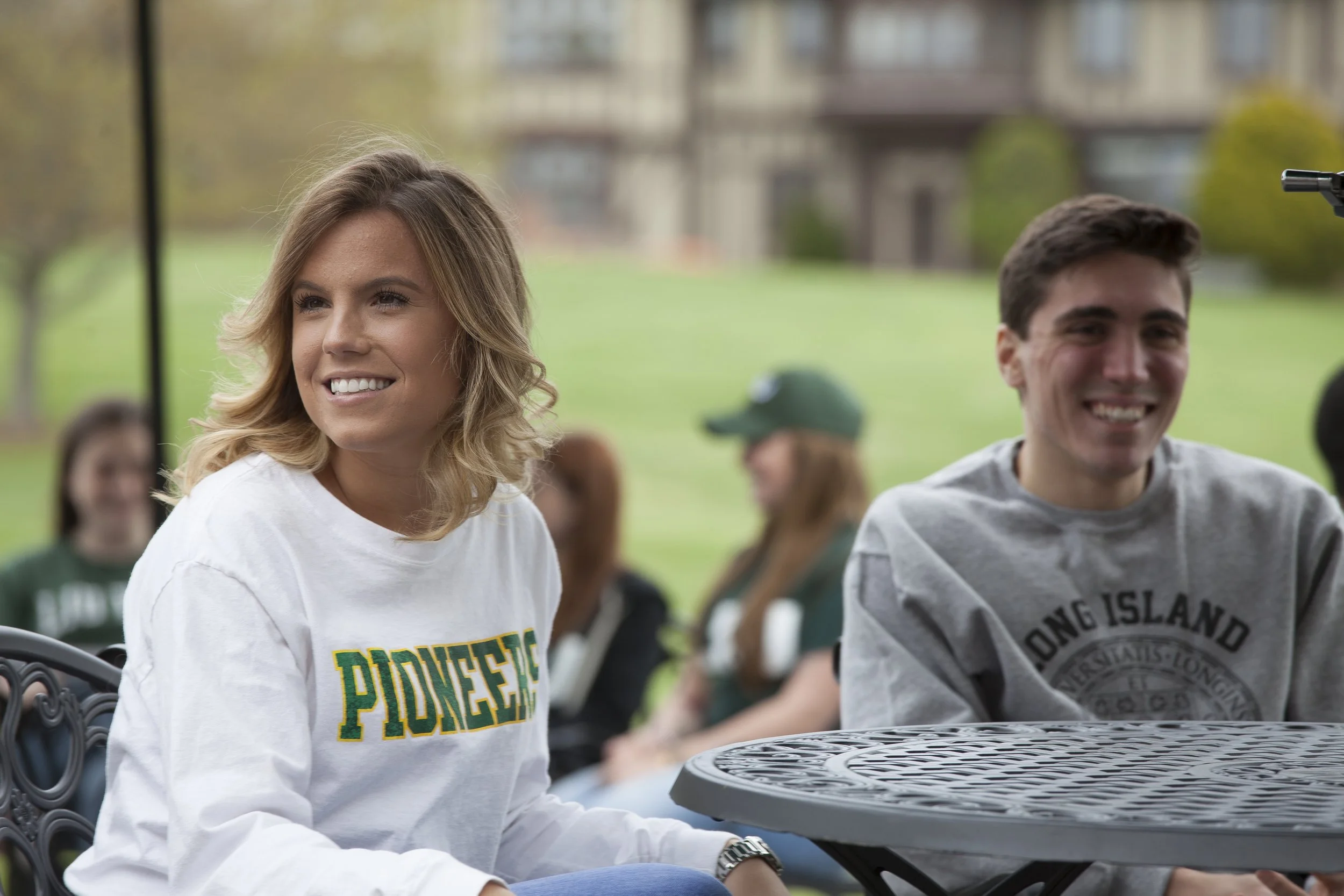 Two young adults, a woman and a man, sitting outdoors at a table, smiling. The woman is wearing a white sweatshirt with 'PIONEERS' written on it, and the man is in a gray sweatshirt with 'LONG ISLAND' on it. In the background, there are other people 