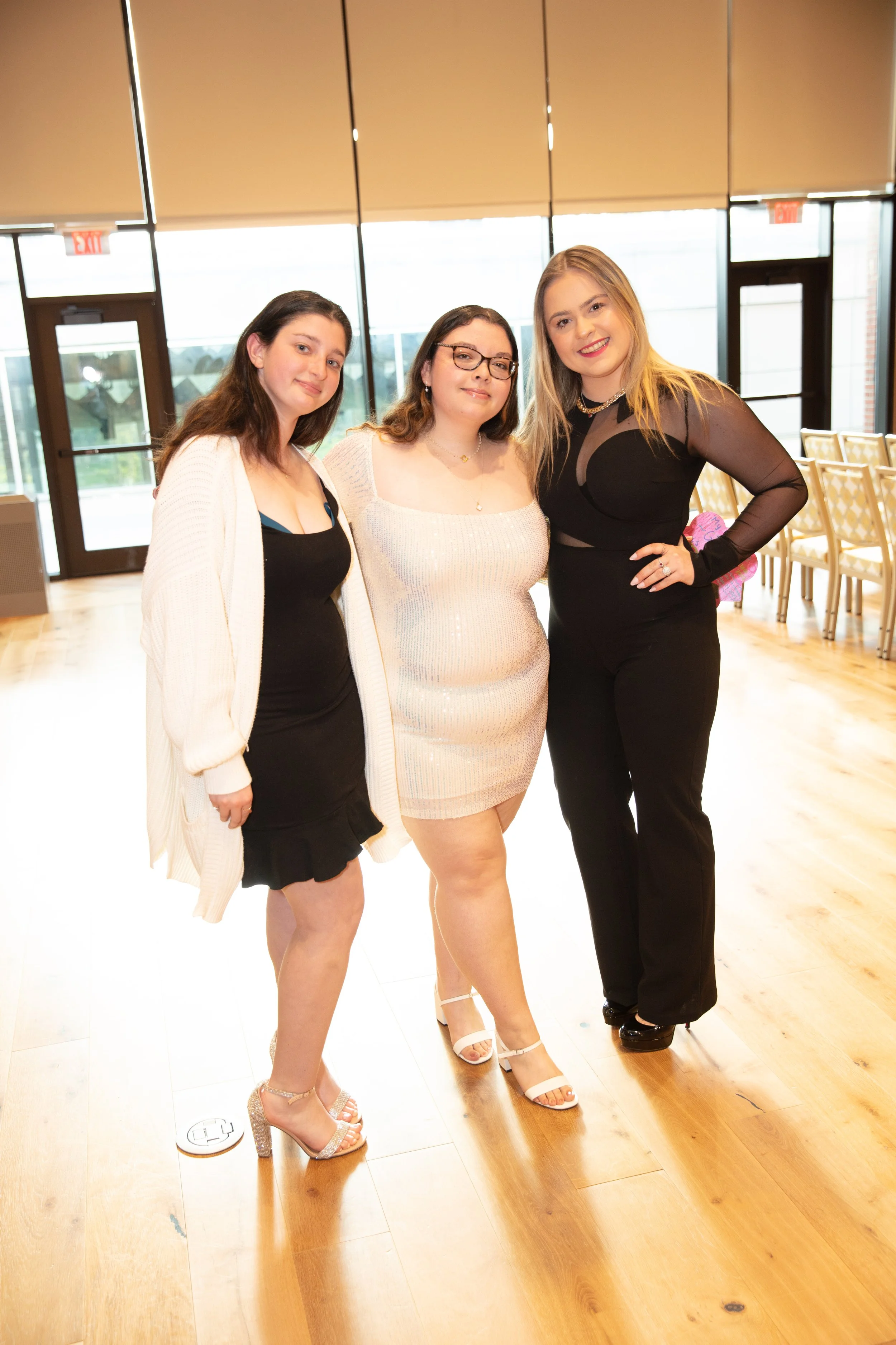 Three women standing close together in a well-lit indoor space with wooden floors, dressed in formal attire, smiling at the camera.