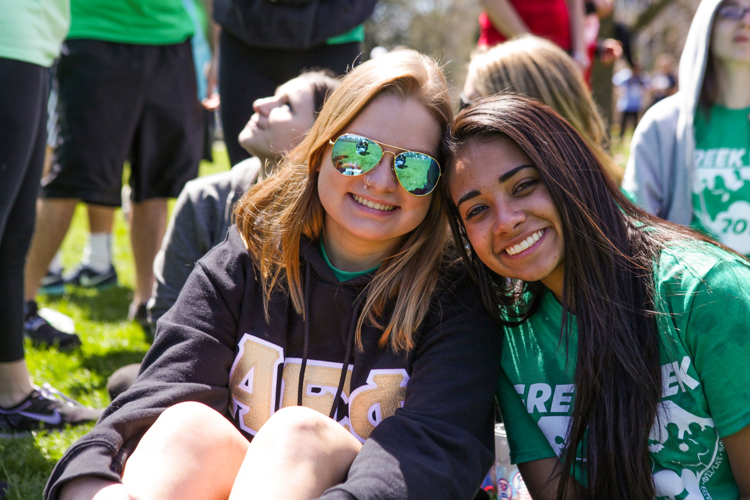 Two smiling young women sitting on grass at an outdoor event, surrounded by other people. One woman has blonde hair, sunglasses, and a black hoodie, while the other has dark hair and a green shirt.