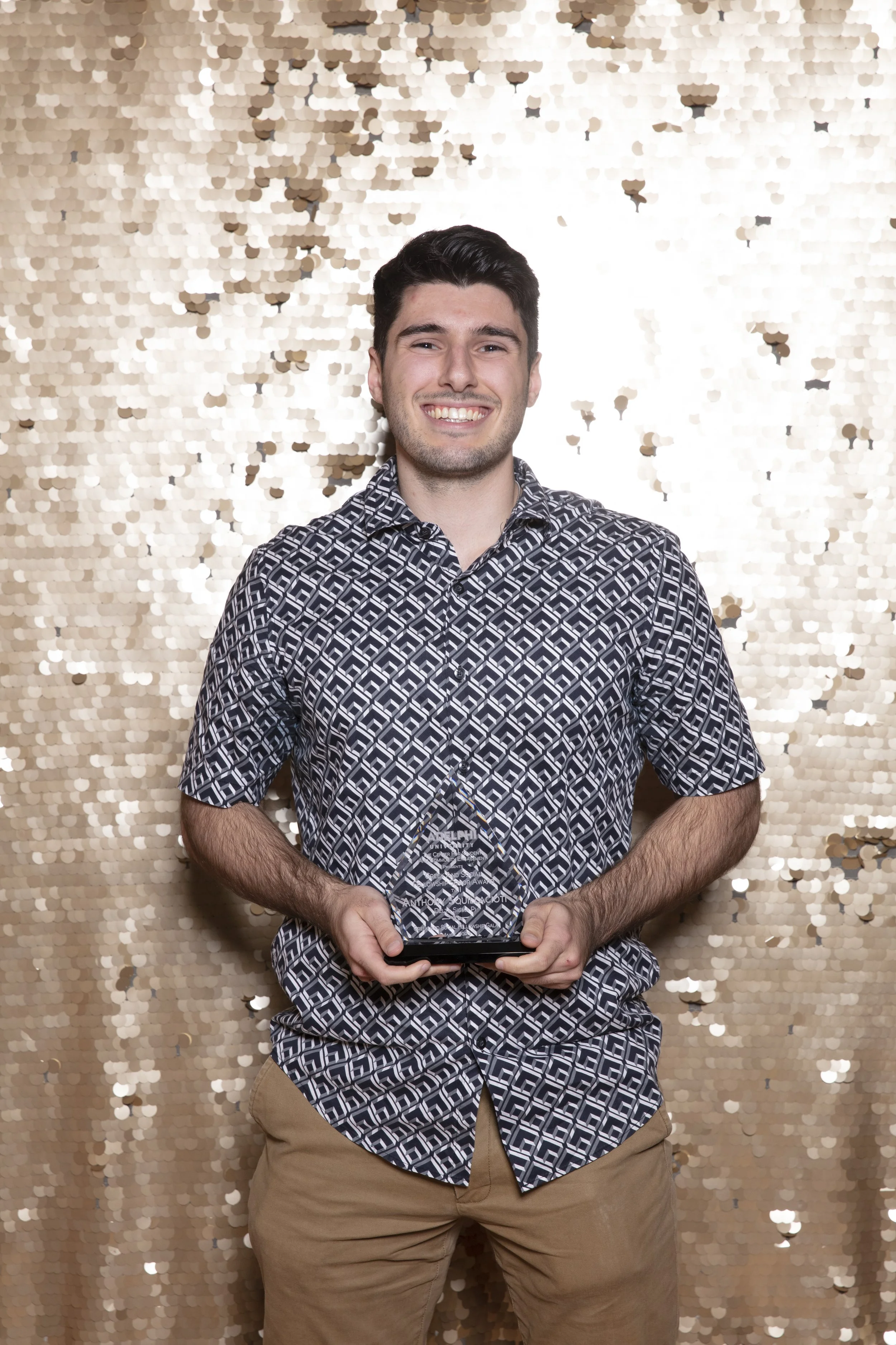 Young man with dark hair and a big smile, wearing a patterned shirt and khaki pants, holding an award plaque, standing against a gold sequin backdrop.