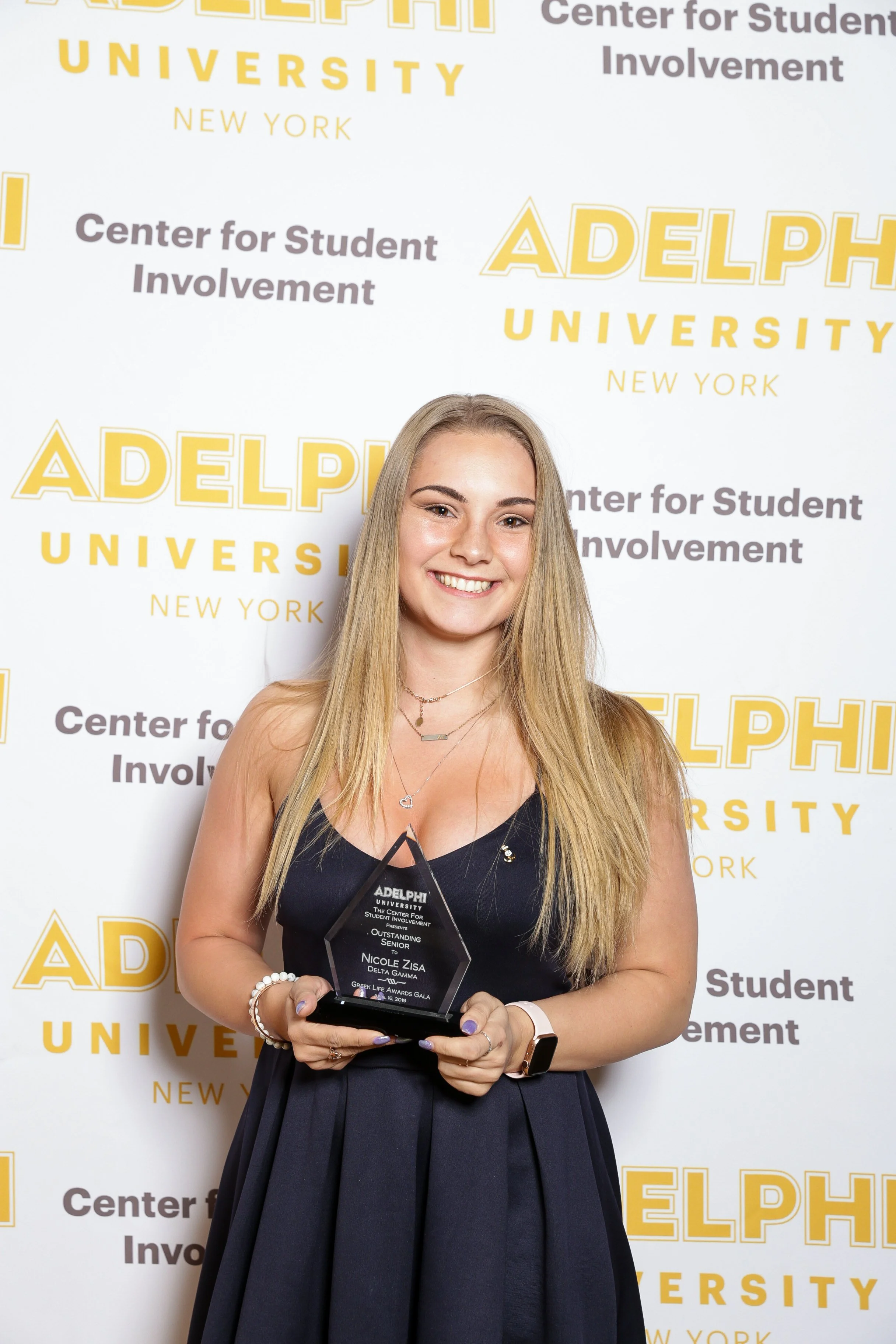 A young woman with long blonde hair smiling and holding an award trophy in front of a backdrop with 'Adelphi University' and 'Center for Student Involvement' written on it.