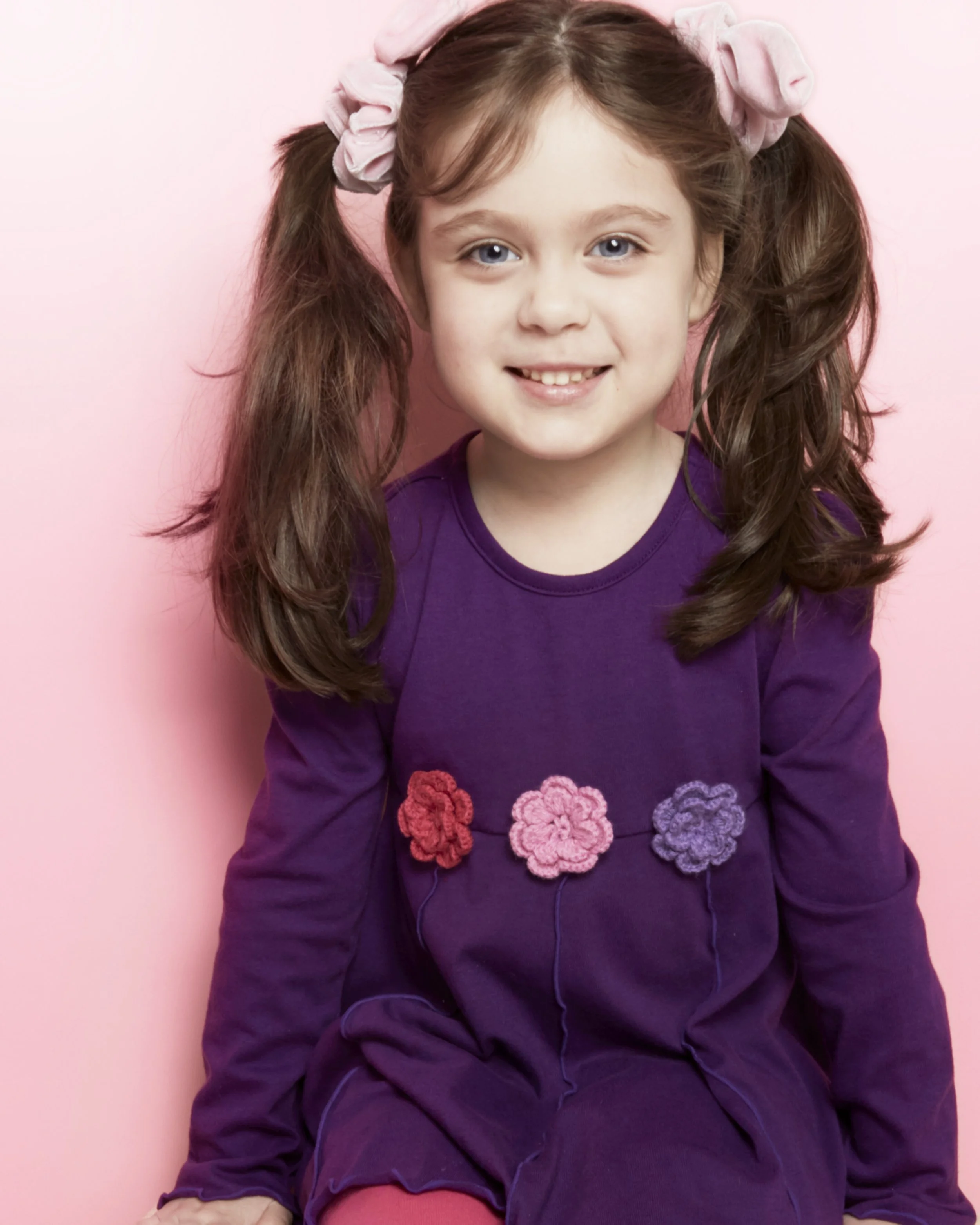 A young girl with brown pigtails tied with pink scrunchies, wearing a purple dress with colorful flower appliqués, smiling and sitting against a pink background.