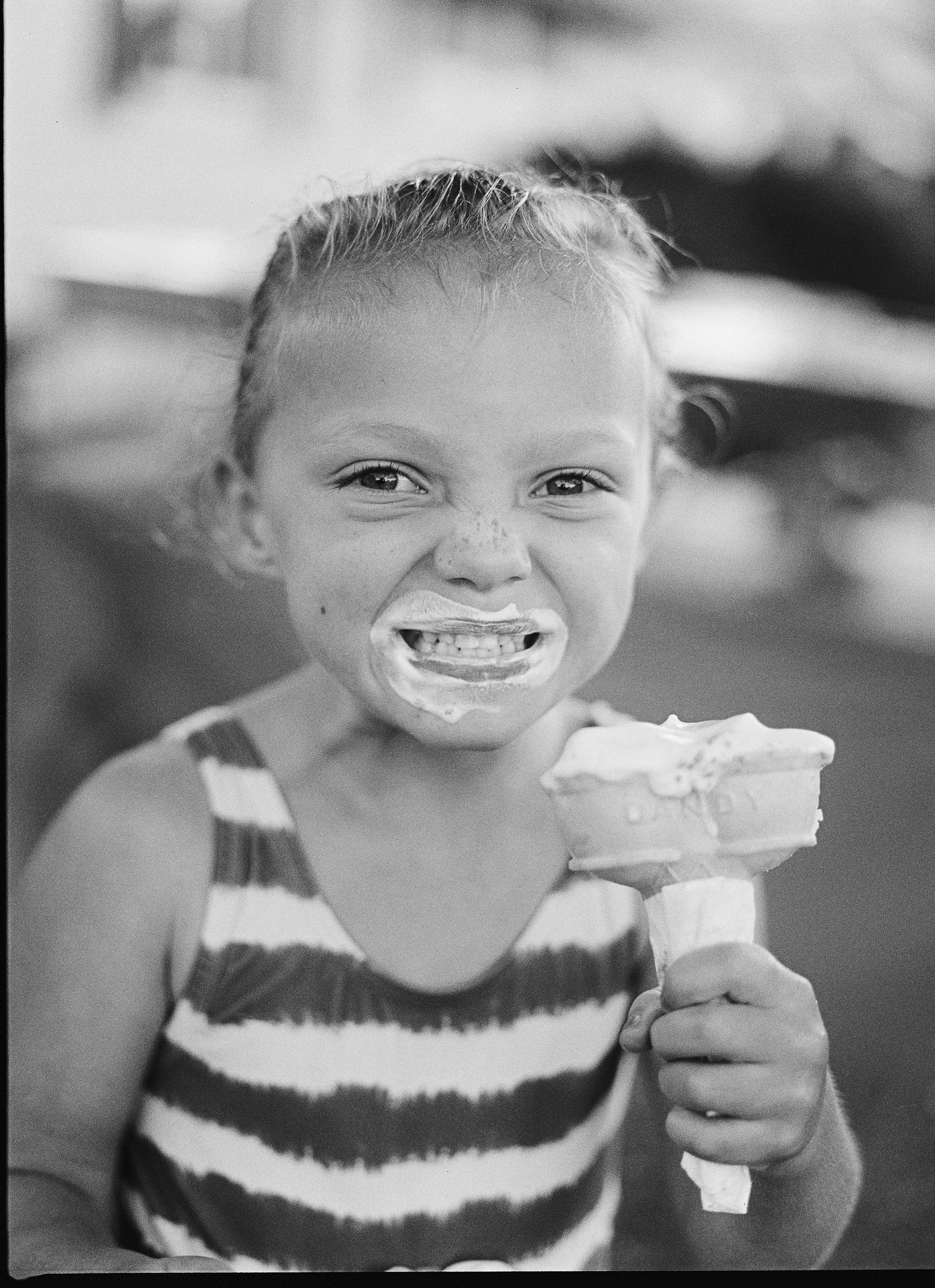 A young girl with a big smile, holding a large ice cream cone, wearing a striped sleeveless top, in black and white photograph.