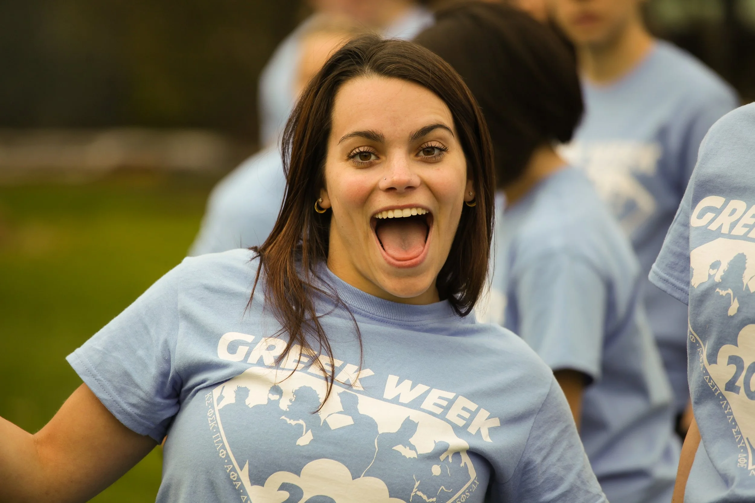 A young woman with dark brown hair, wearing hoop earrings, smiling with her mouth open, wearing a light blue T-shirt with the words "Great Week" and a design showing mountains and clouds, outdoors with other people in similar T-shirts in the backgrou