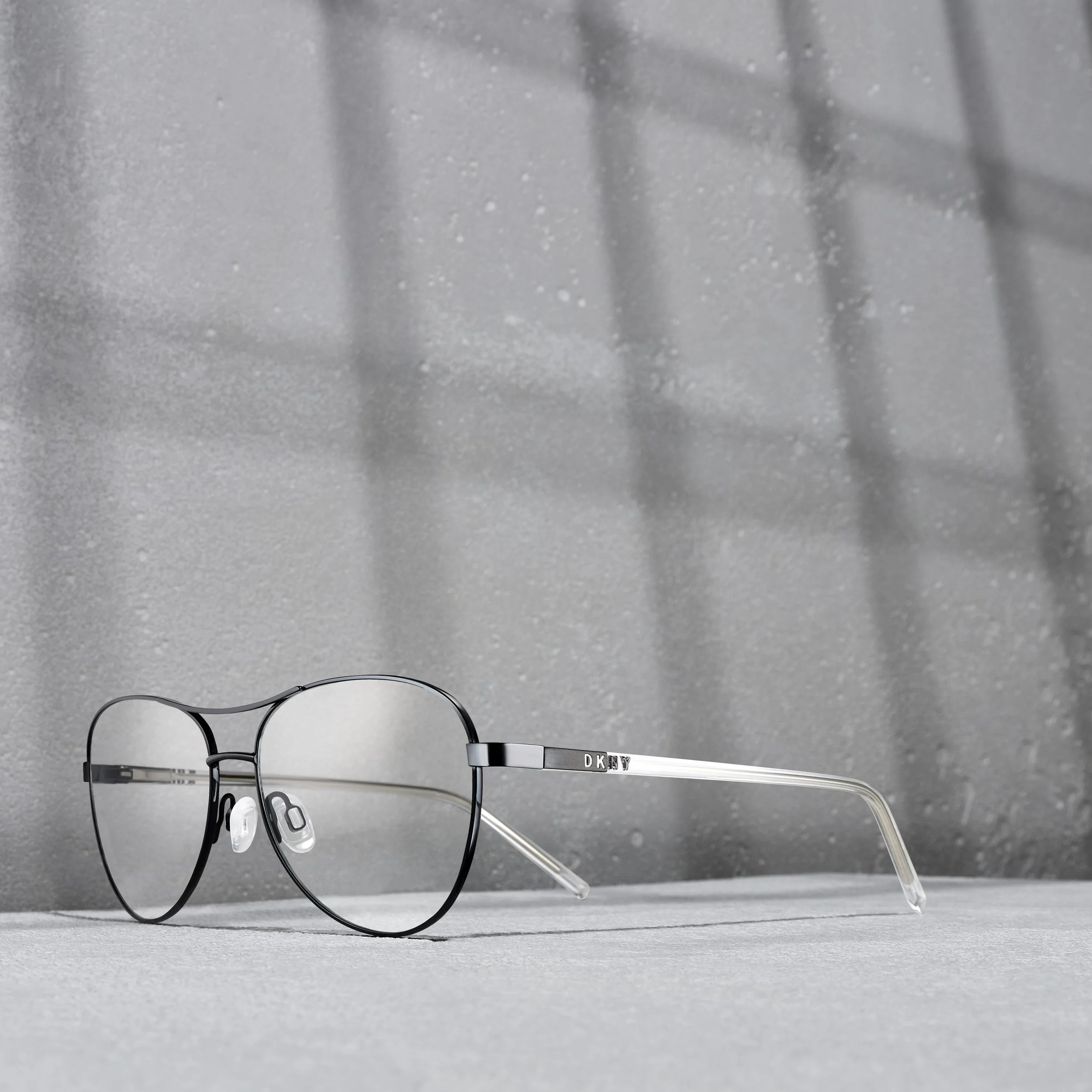 Black-framed eyeglasses with clear temples resting on a gray surface, with a textured gray background and shadow reflections.