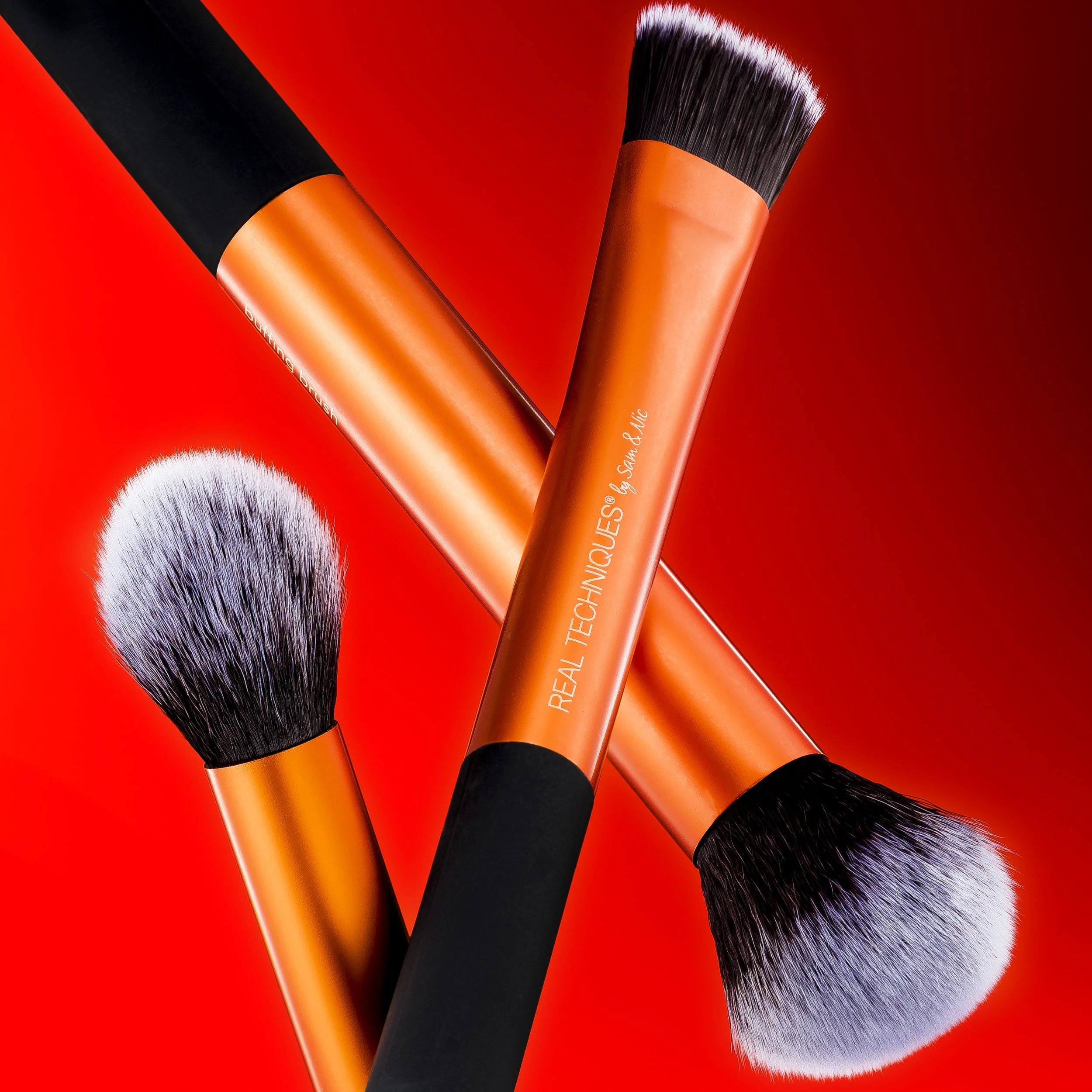 Three makeup brushes with orange handles, black ferrules, and white-to-black ombre bristles, arranged on a red background.