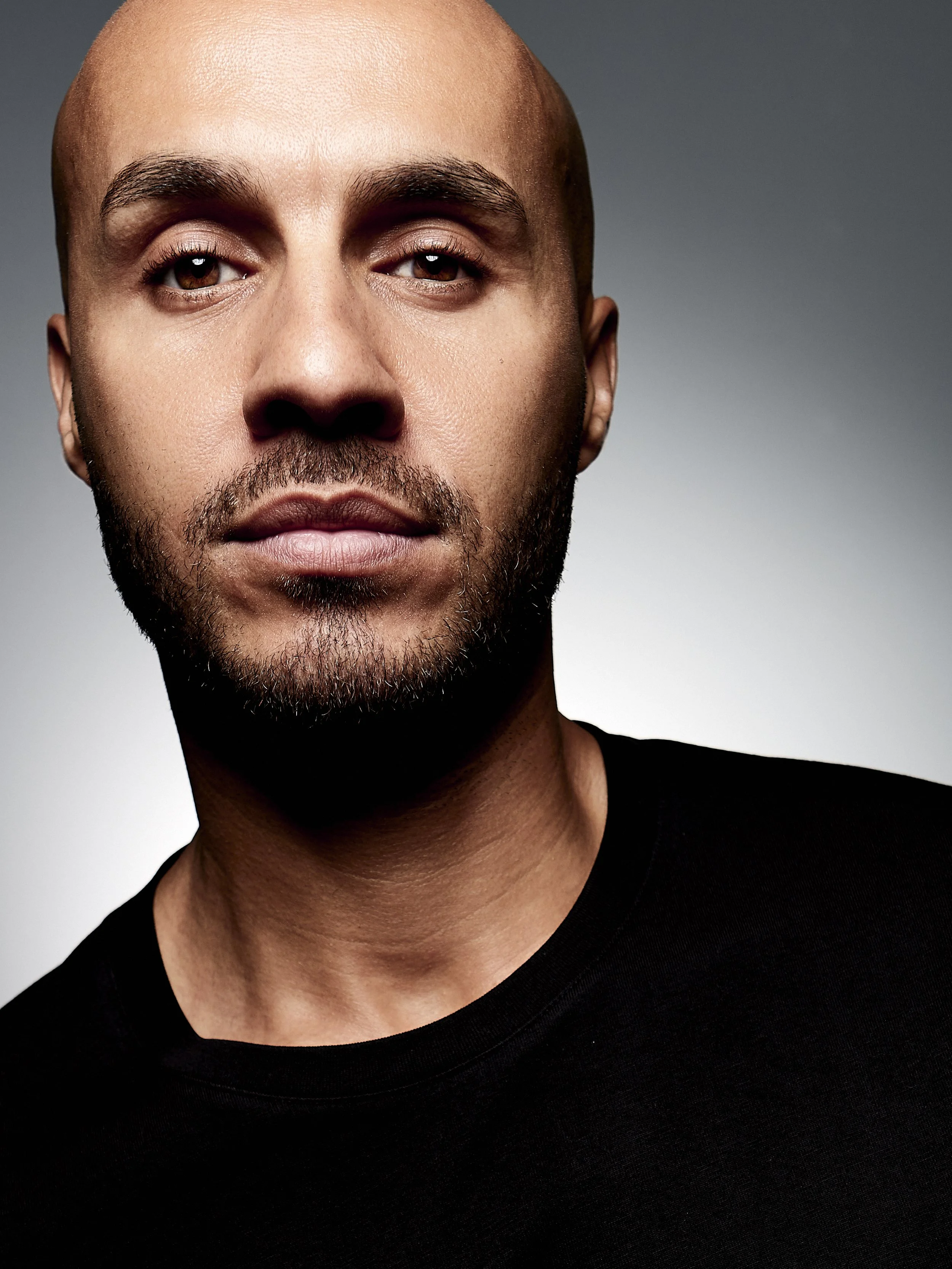 Close-up portrait of a bald man with a beard and brown eyes, wearing a black shirt, against a gray background.