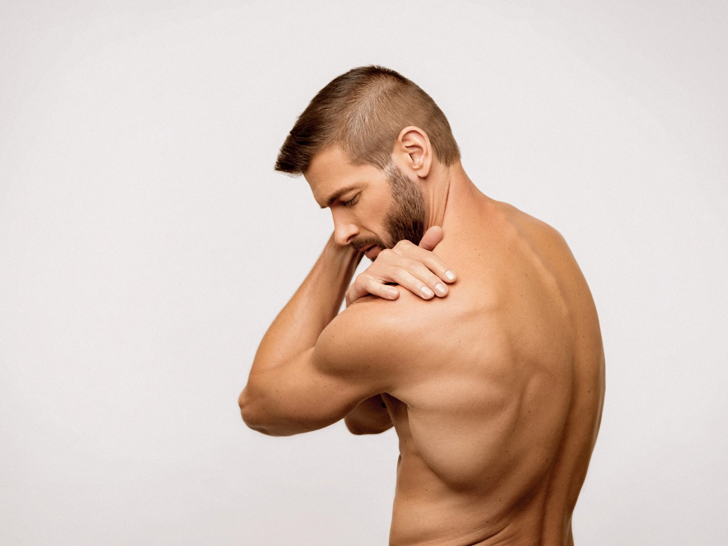 A shirtless man with a beard, holding his shoulder and looking down, standing against a plain white background.