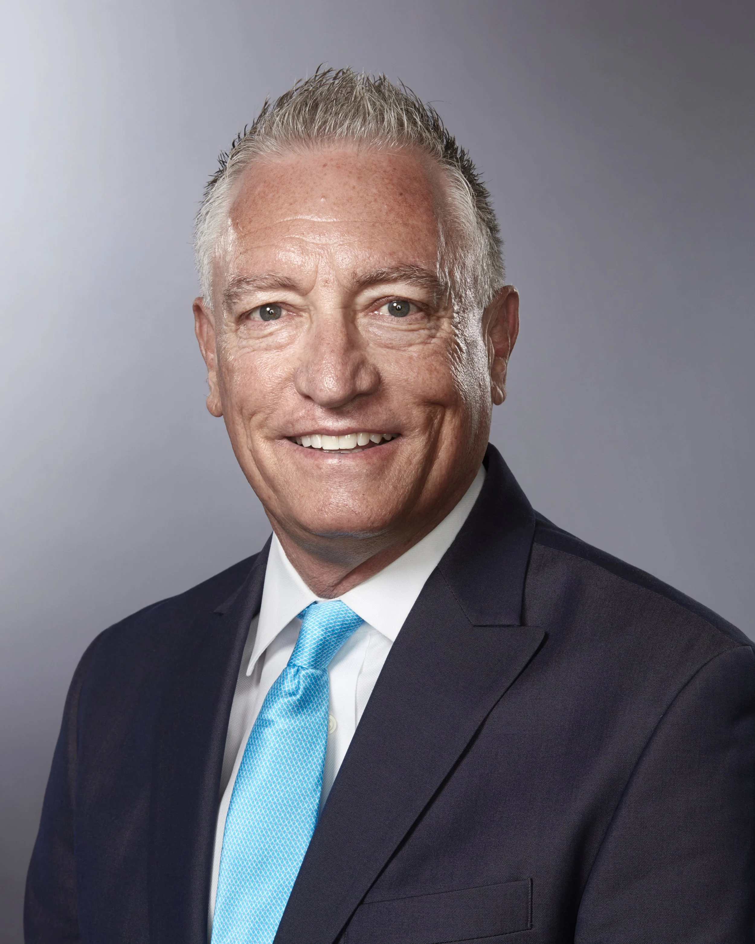 A professional headshot of a middle-aged man with short, gray hair, wearing a dark suit, white shirt, and a light blue tie, smiling against a neutral gray background.