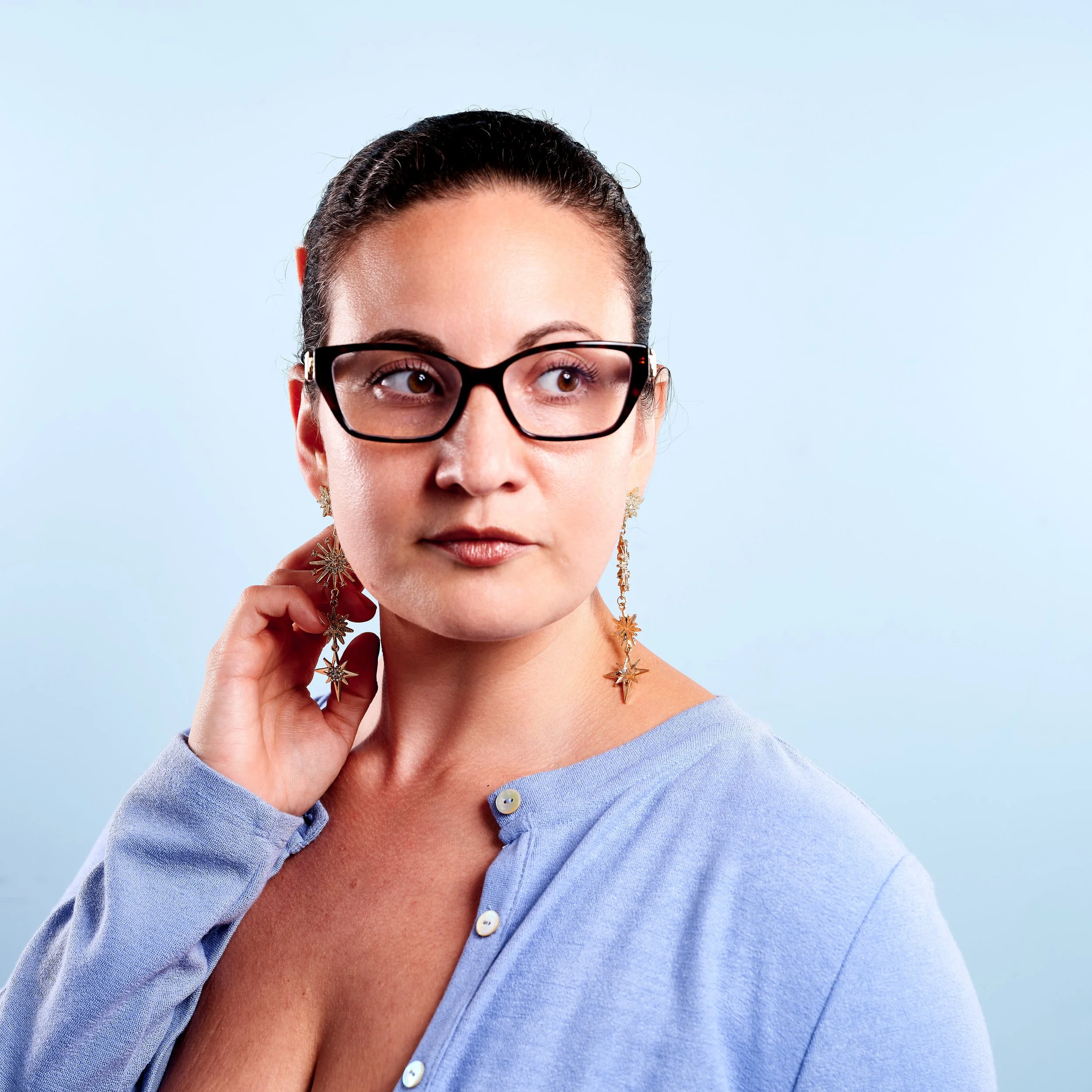 Close-up of a woman with dark hair, wearing glasses, jewelry, and a blue shirt, against a light blue background.