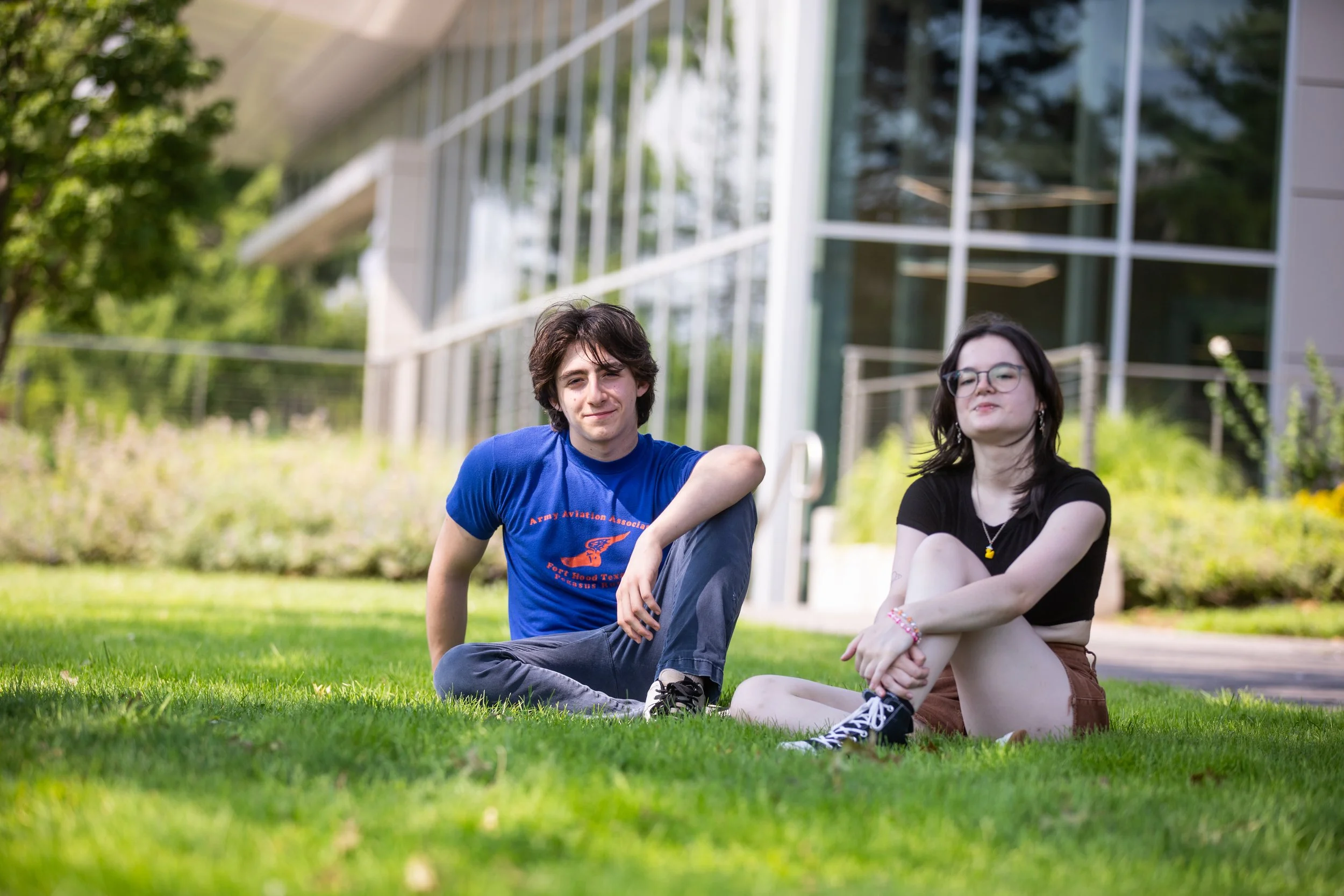 A young man and young woman sitting on a grassy lawn outside a modern building, smiling and relaxing.