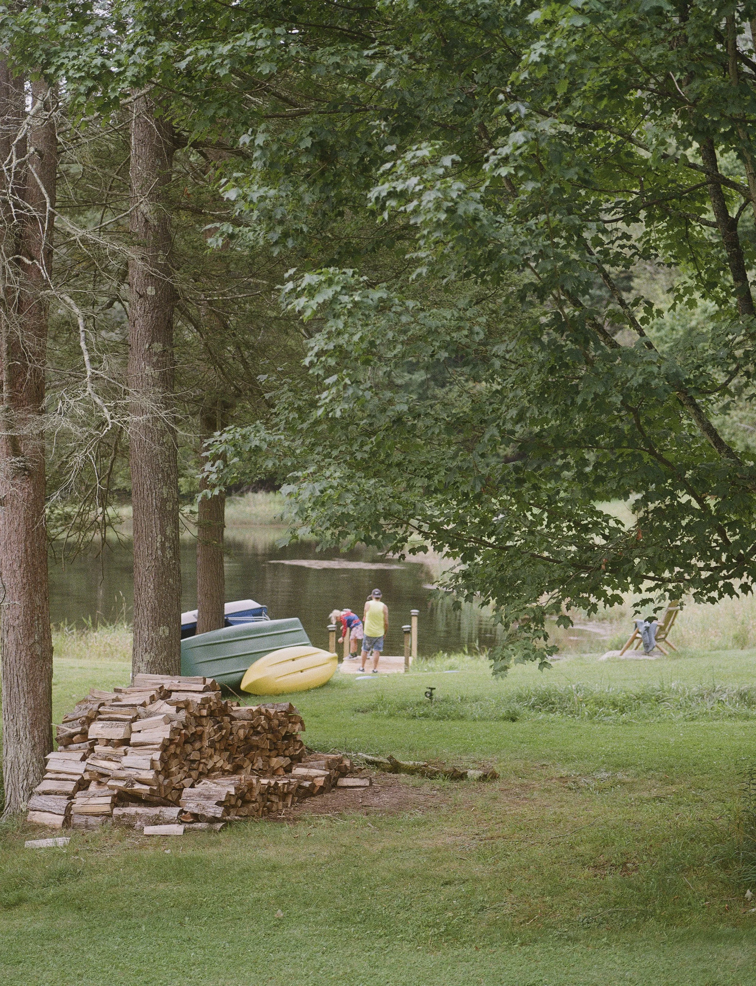 A grassy area with three large trees near a body of water. A stack of firewood, two kayaks, and a yellow boat are on the grass. Two people are near the water, one wearing a hat and a green shirt, the other a red shirt. A chair is on the right side of