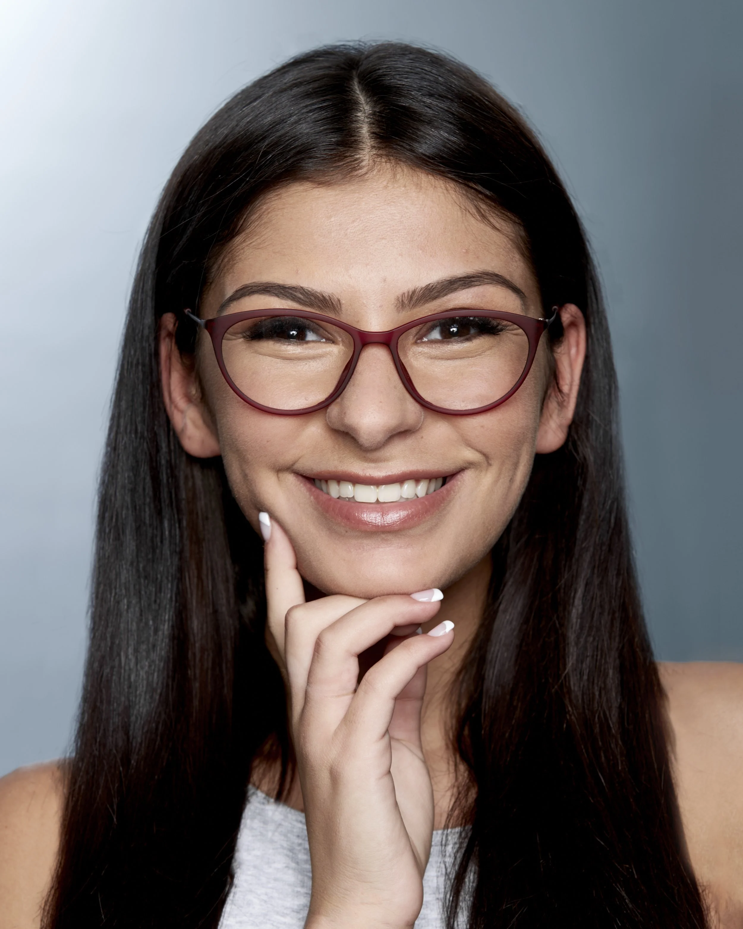 A young woman with long dark hair, wearing glasses, smiling and touching her chin.