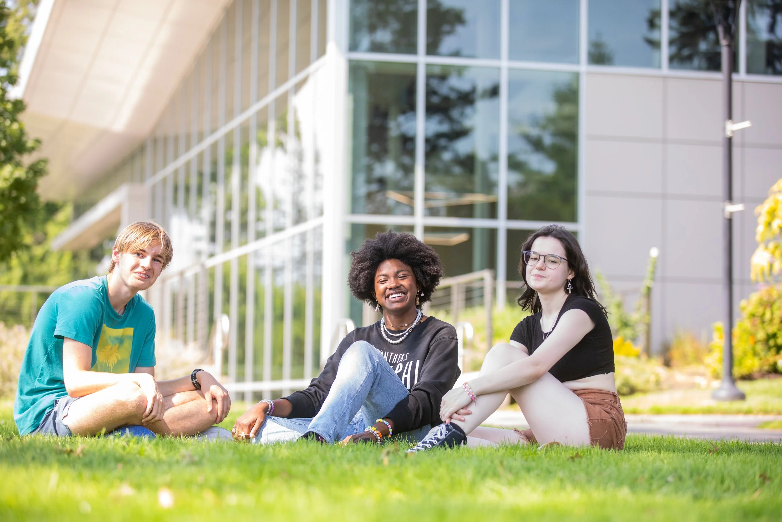 Three young people sitting on the grass in front of a modern glass building, smiling and posing for the photo, with trees and bushes in the background.