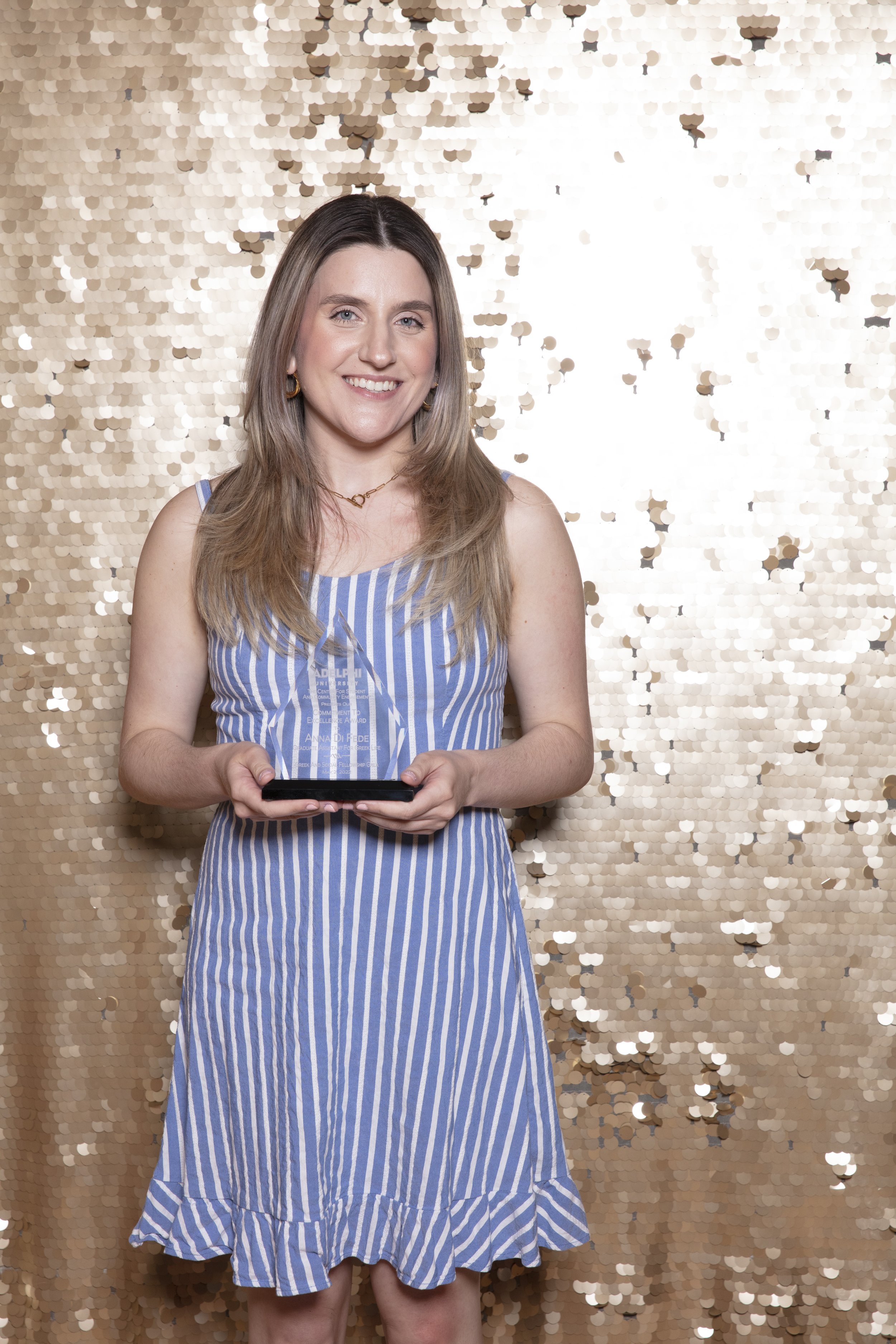 A woman with long blonde hair smiling and holding a glass award, standing in front of a gold sequin backdrop, wearing a blue and white striped sleeveless dress.