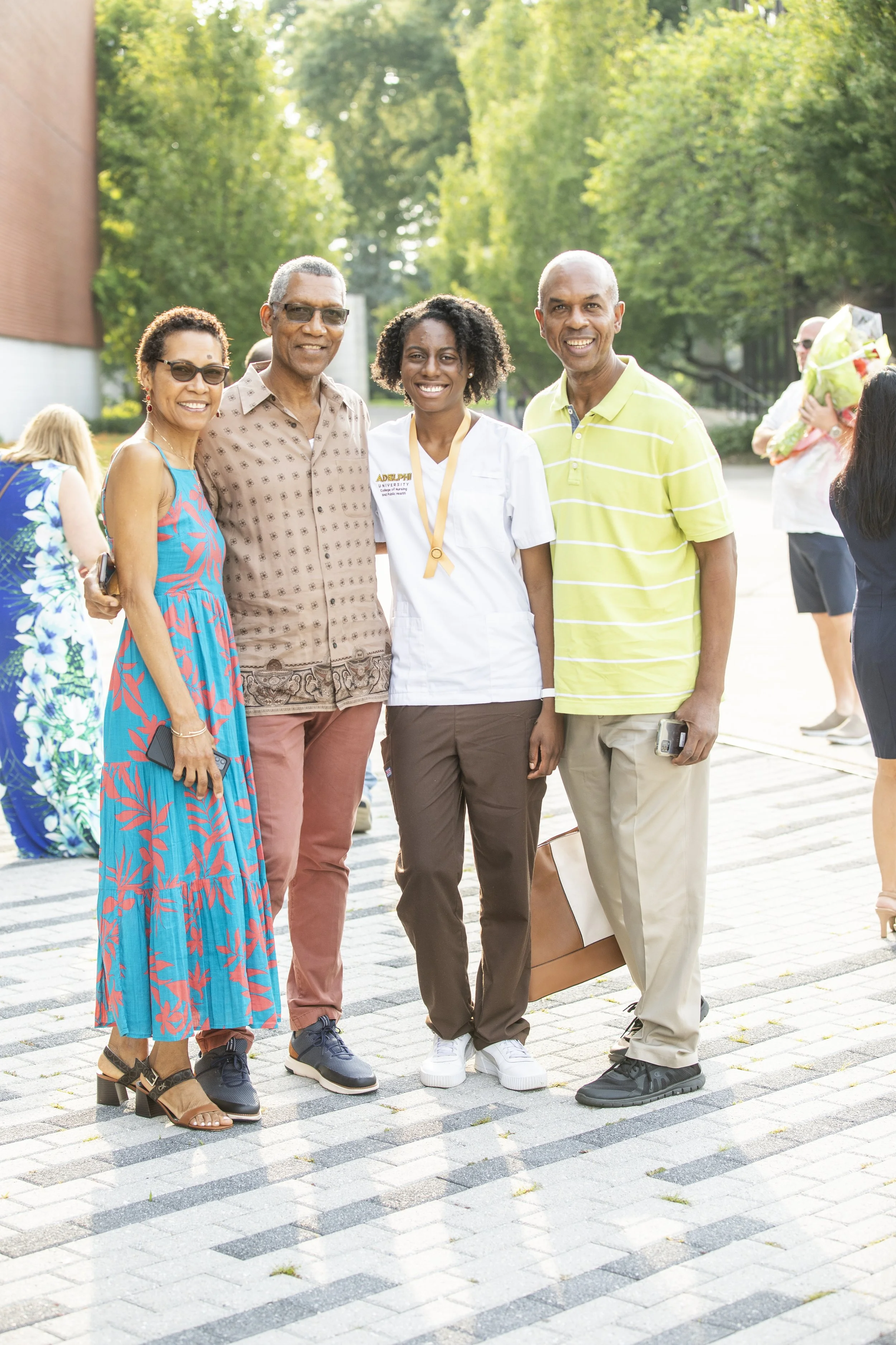 Group of five people standing together outdoors, celebrating a graduation. One person in the center is wearing a white medical uniform and a medal, indicating a nursing or medical degree. Other individuals are dressed casually, smiling, and standing 