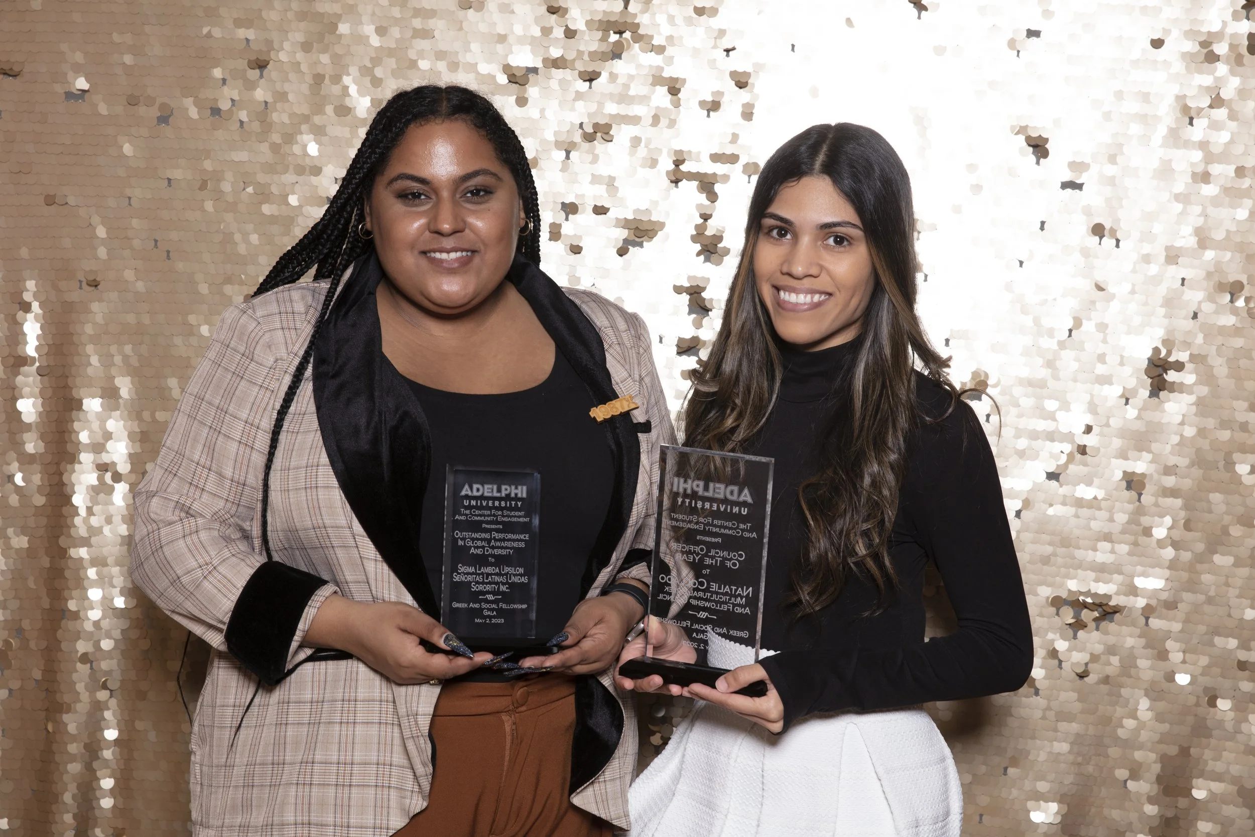 Two women standing together, each holding a clear glass award, smiling in front of a gold sequin backdrop.