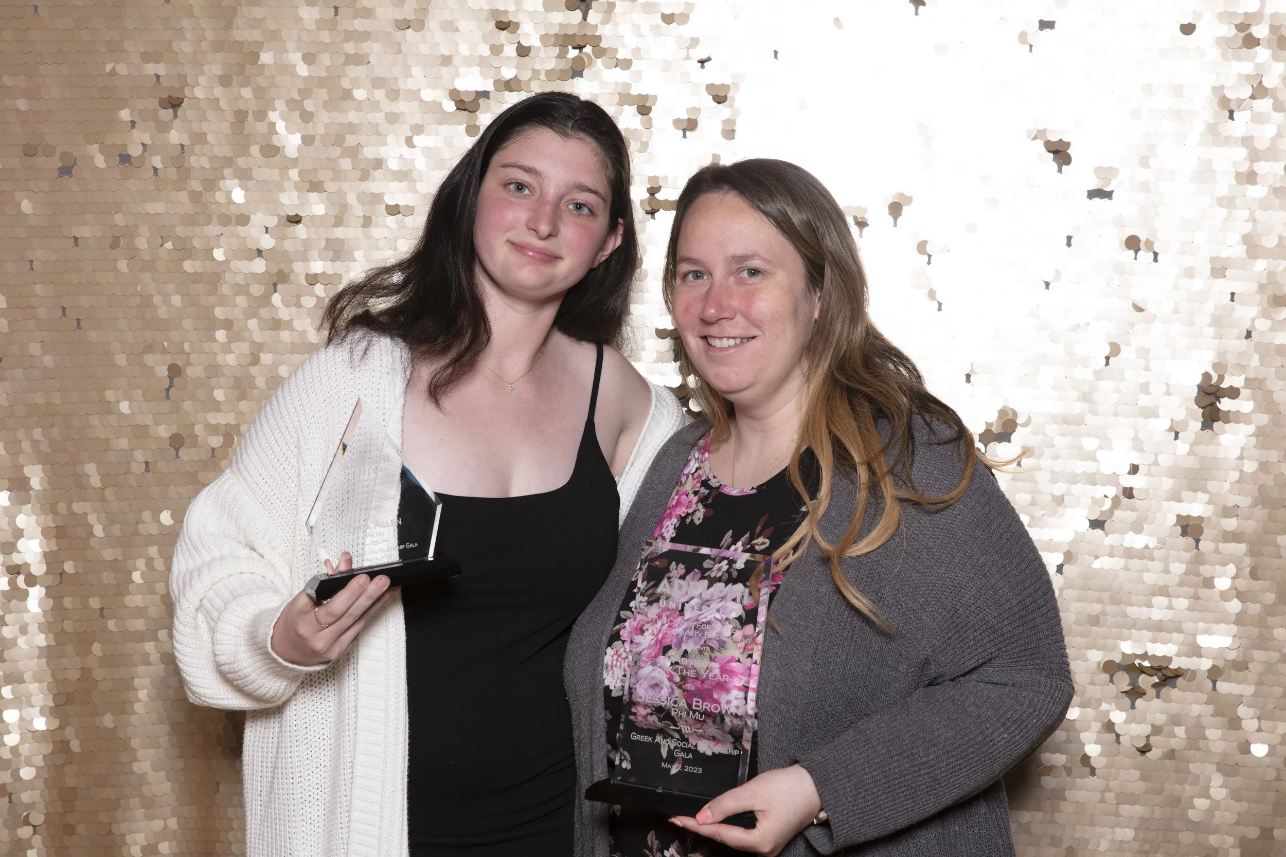 Two women at an awards ceremony holding glass awards, standing in front of a gold sequin backdrop.
