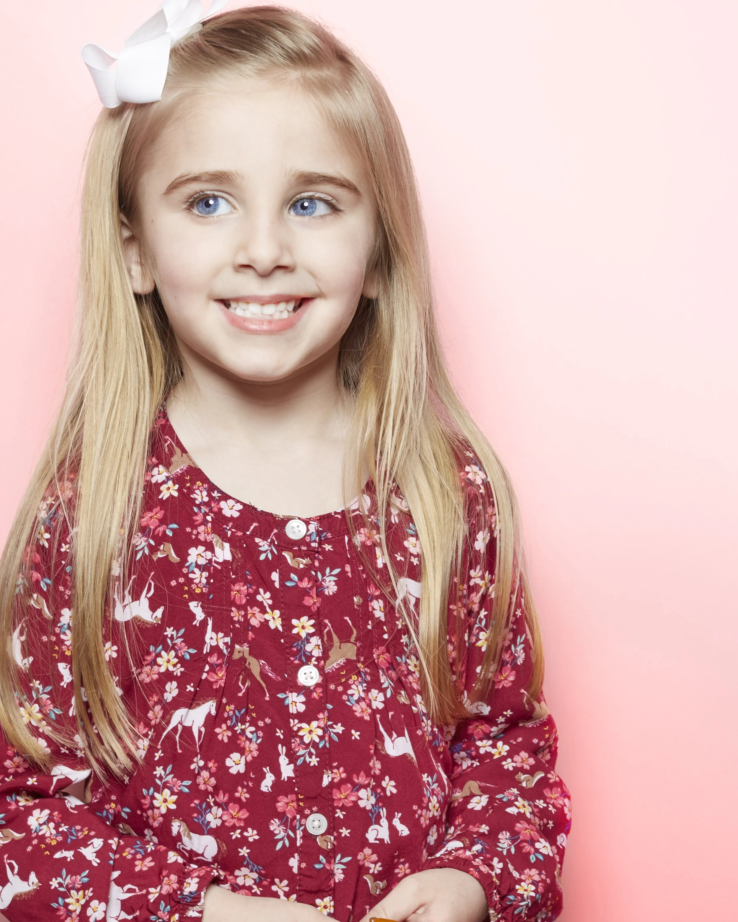 A young girl with long blonde hair wearing a red dress with animal and floral patterns, and a white bow in her hair, smiling against a pink background.