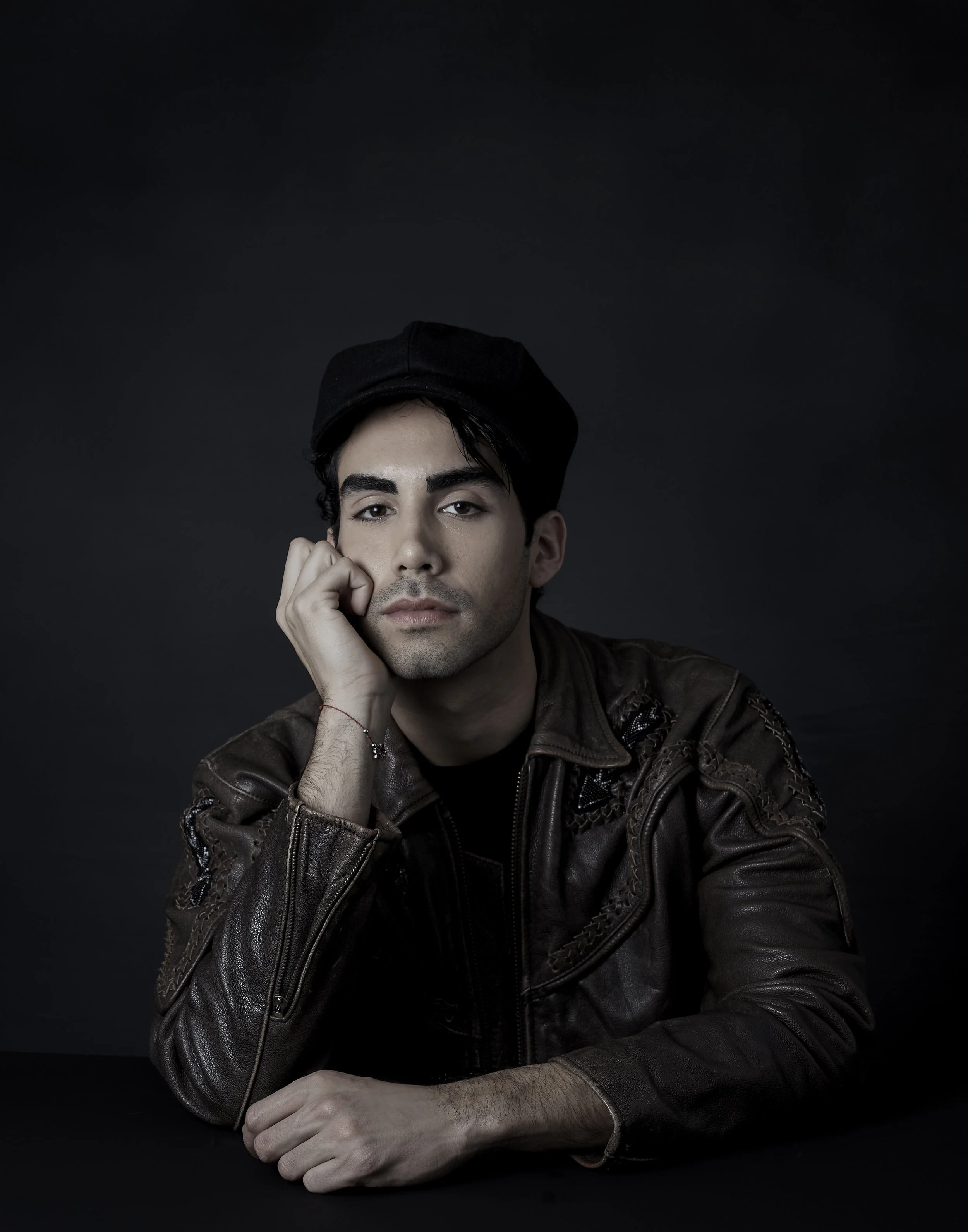 Portrait of a young man with dark hair, wearing a black cap and brown leather jacket, resting his head on his hand against a dark background.