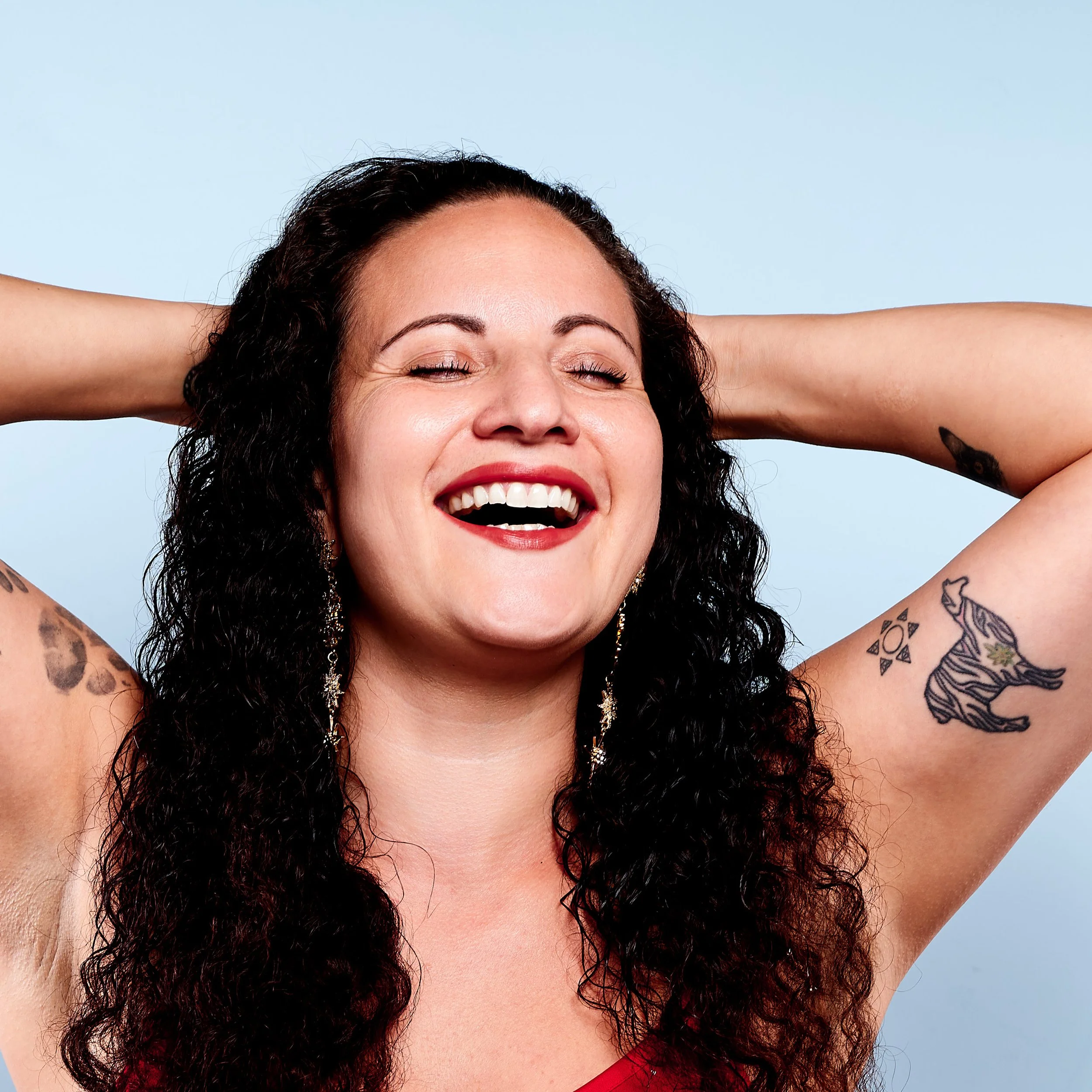 A woman with curly dark hair smiling with her eyes closed, wearing a red top, with arms raised behind her head, against a light blue background.