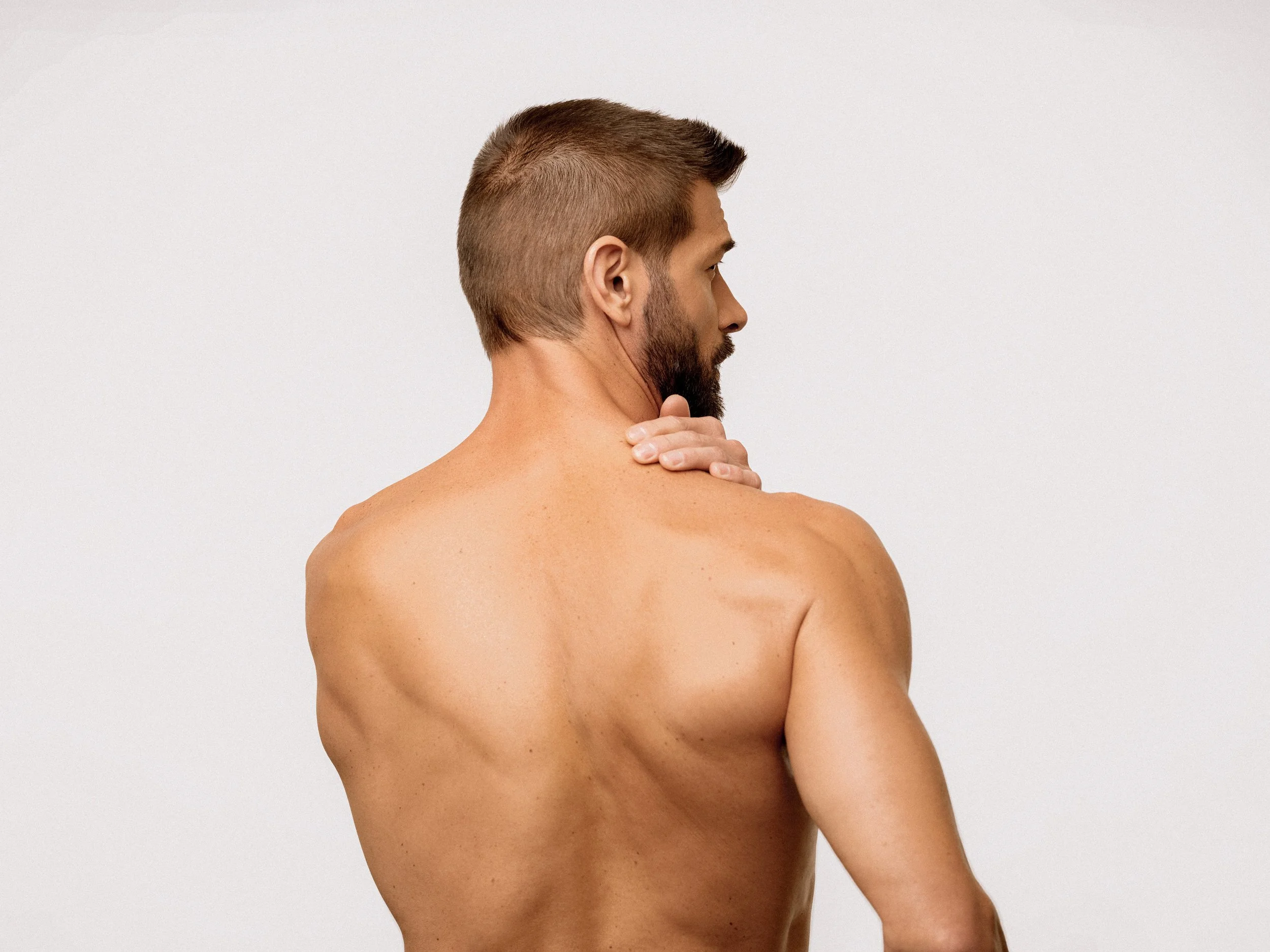 A shirtless man with short brown hair and a beard, seen from the back, touching his neck with one hand against a plain light gray background.
